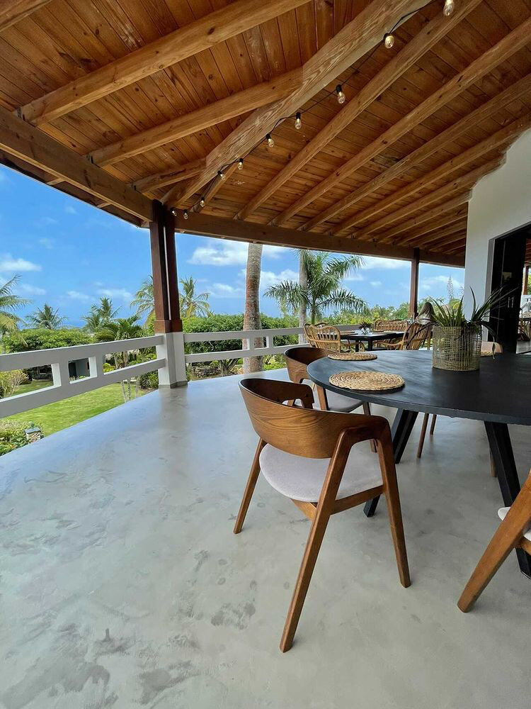 Outdoor patio with wooden ceiling, modern chairs around a dark table, string lights, and a view of palm trees and a garden.