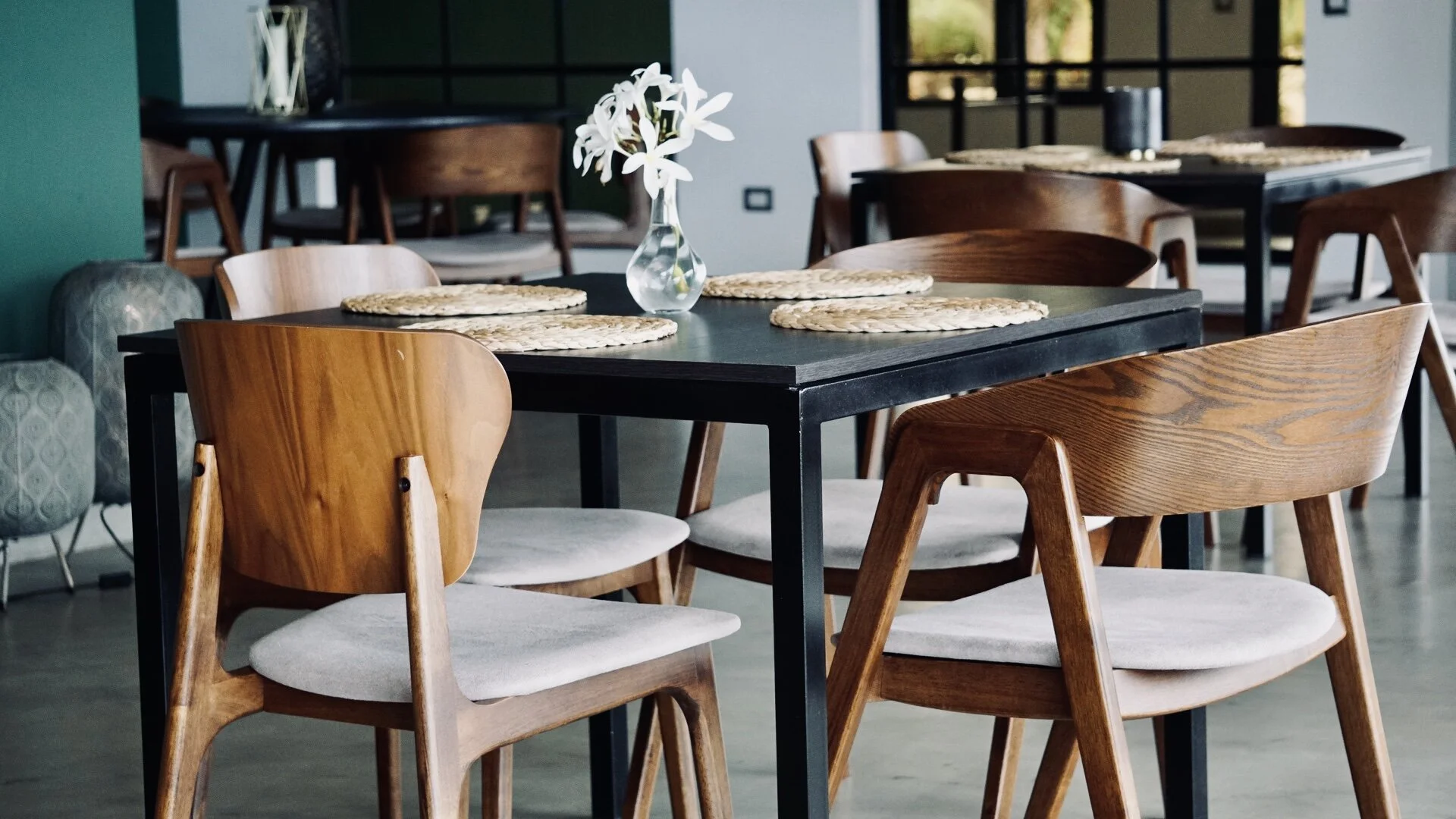 Modern dining area with wooden chairs and a black table, adorned with woven placemats and a vase of white flowers.