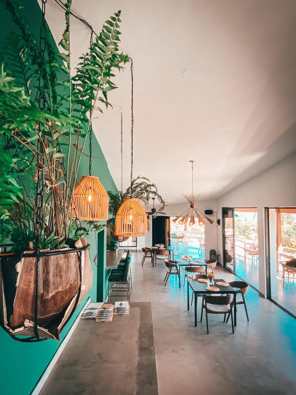 Modern restaurant interior with green walls, hanging plants, and wooden furniture. The space includes tables, chairs, pendant lights, and large windows with outdoor views.
