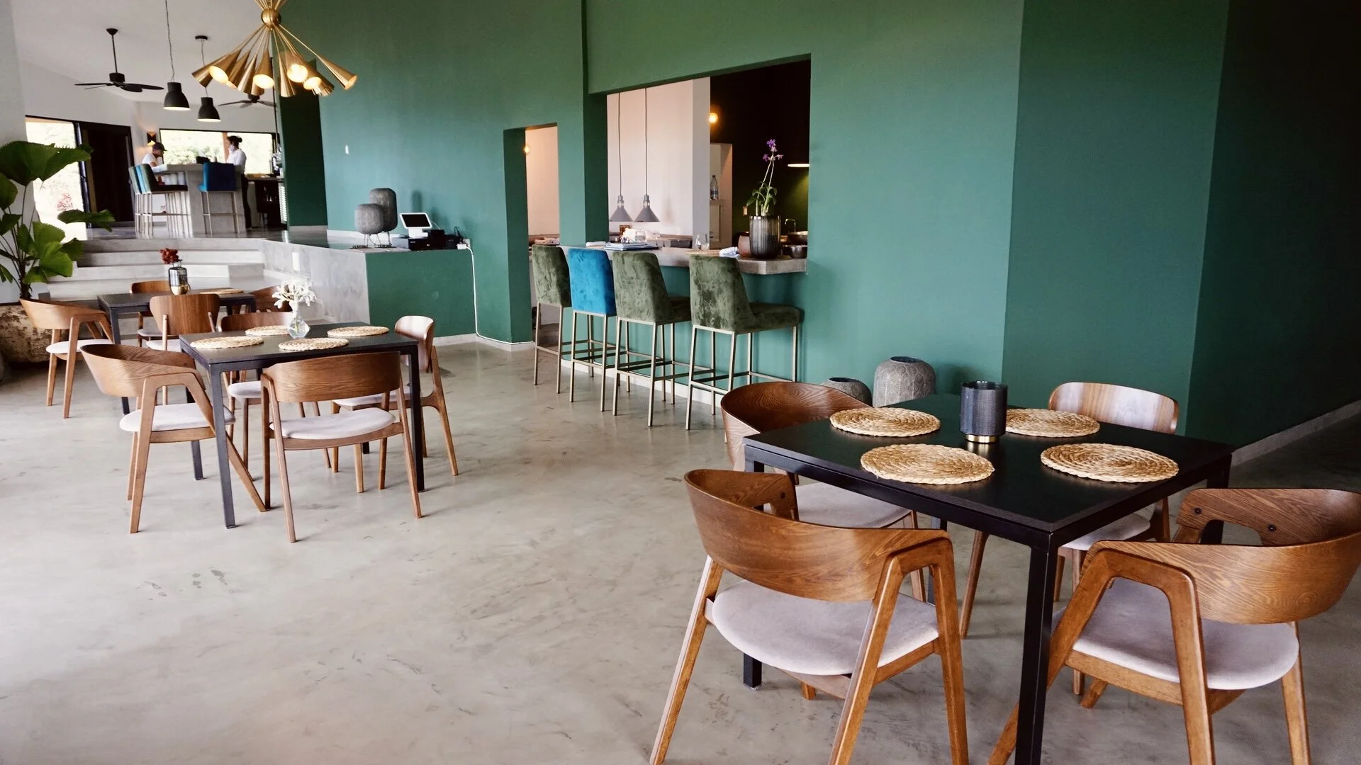 Modern restaurant interior with wooden tables and chairs, green walls, and decorative pendant lighting.