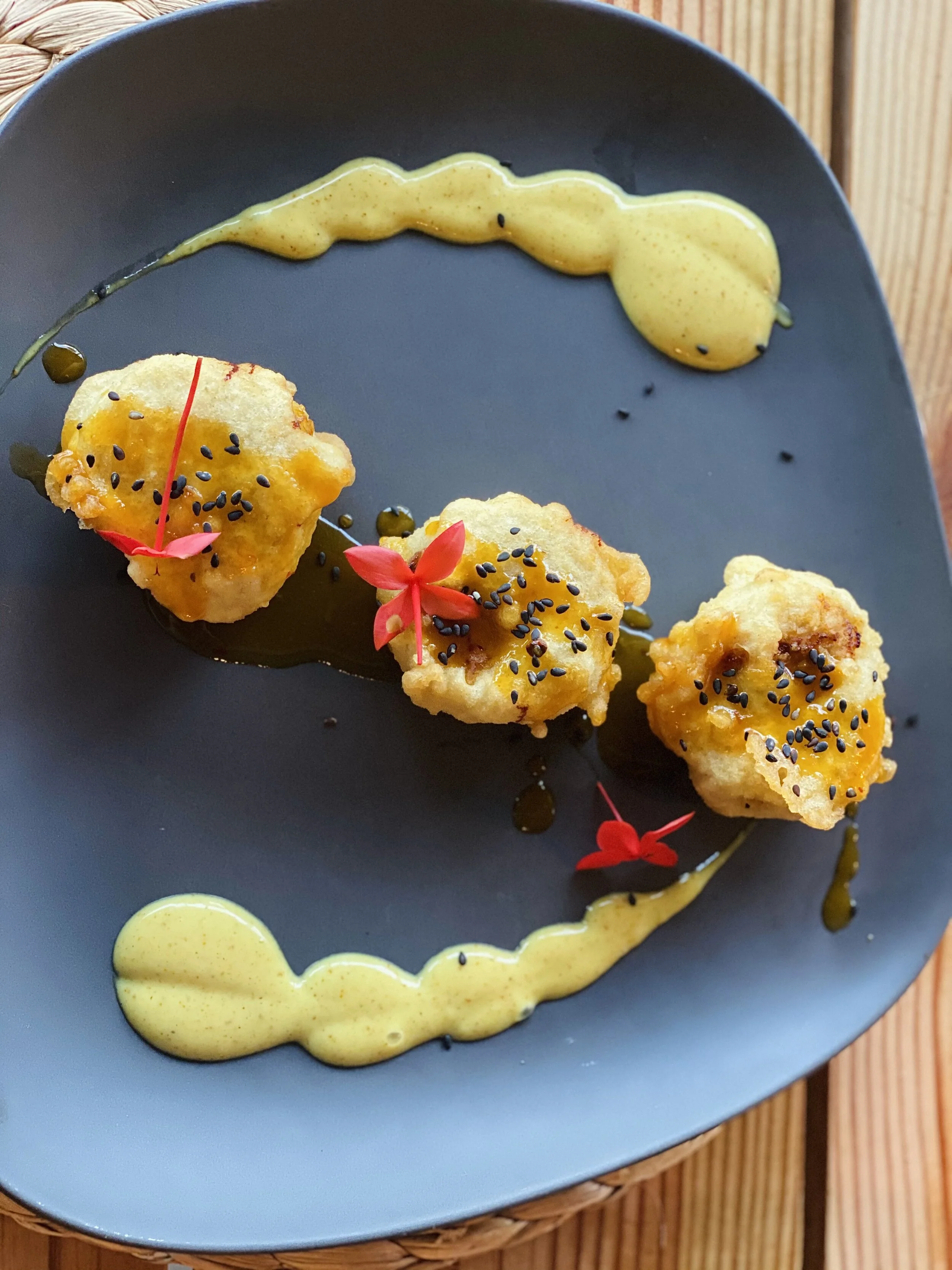 Plate of three small, golden-brown fried items garnished with black sesame seeds and red petals, served with a drizzle of yellow sauce on a dark plate.