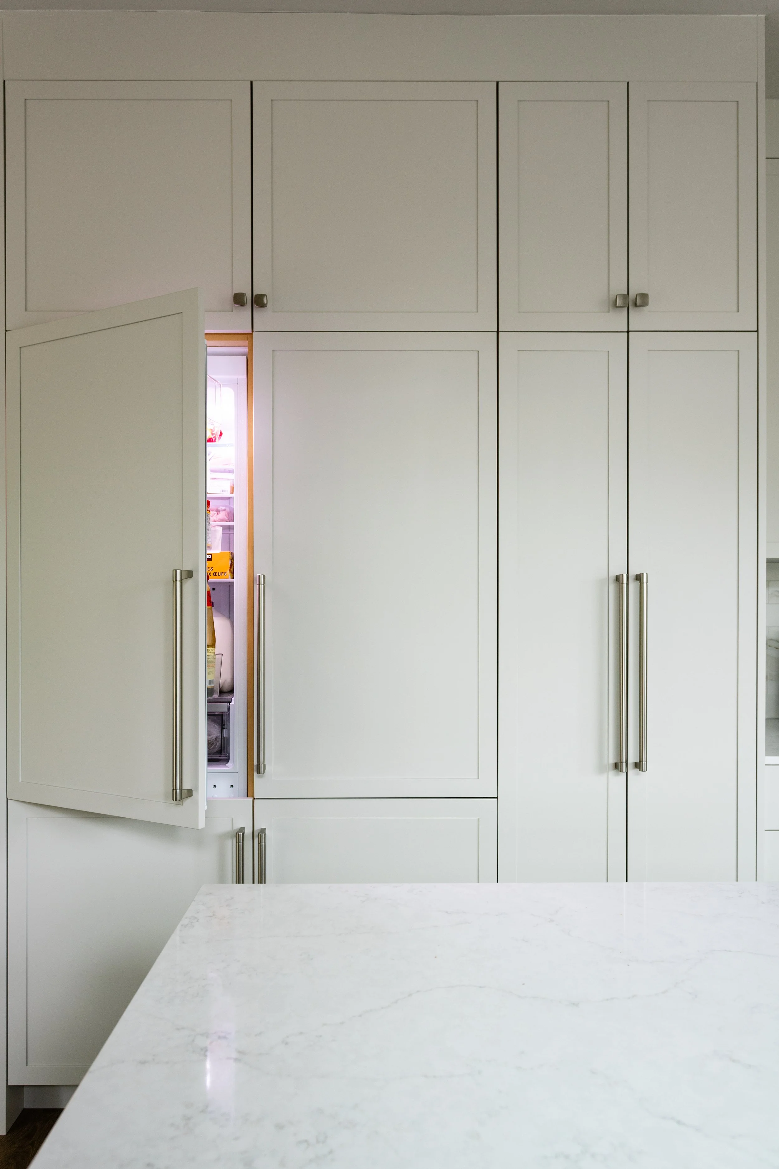 Open refrigerator door behind white kitchen cabinets and a white marble kitchen island.