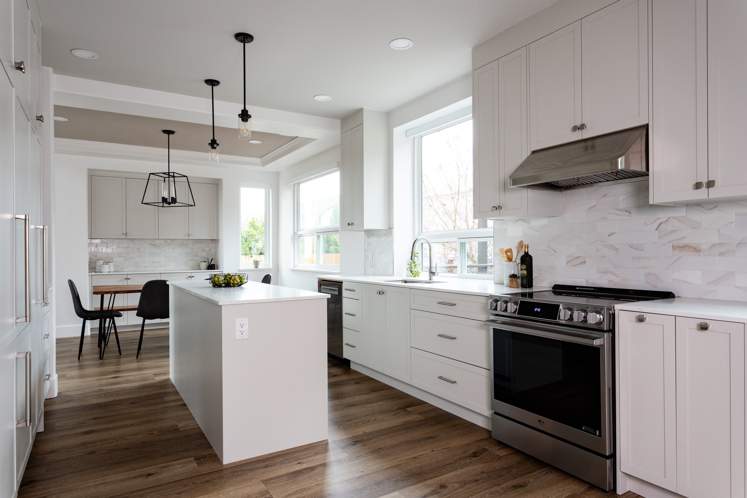 Modern kitchen with white cabinets, marble backsplash, stainless steel oven, island with fruit bowl, and large windows letting in natural light.