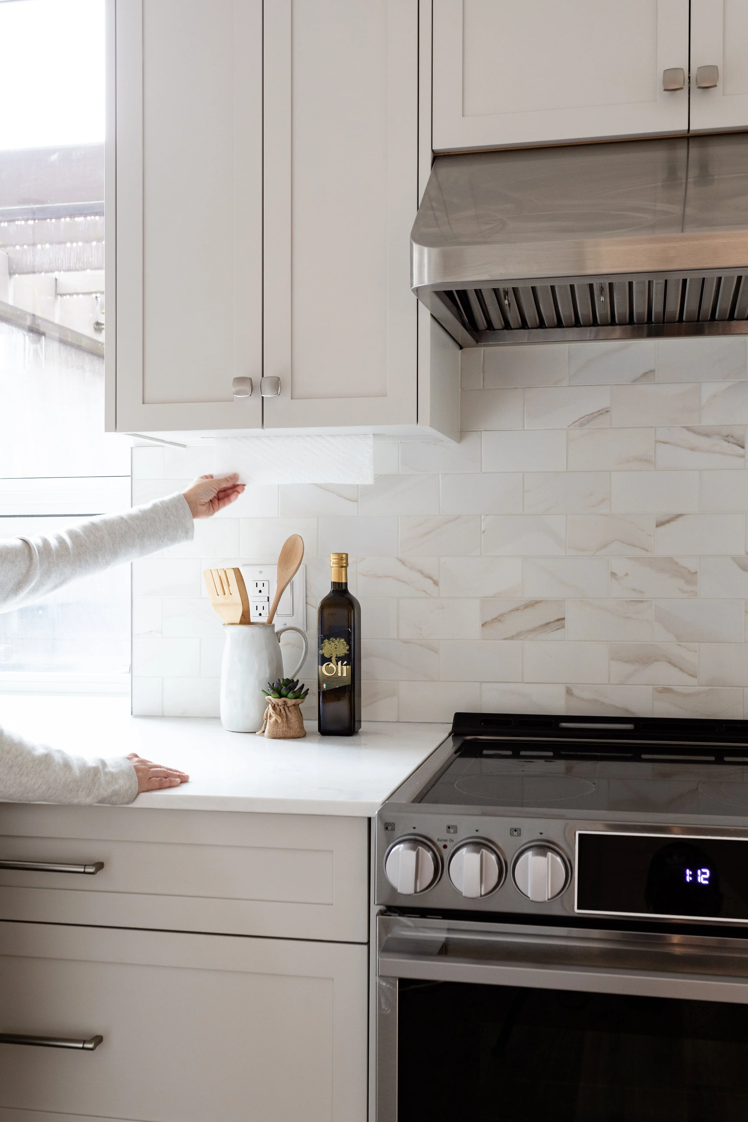 Person reaching for paper towel roll above kitchen counter with stove, cabinets, and decorative items in the background.