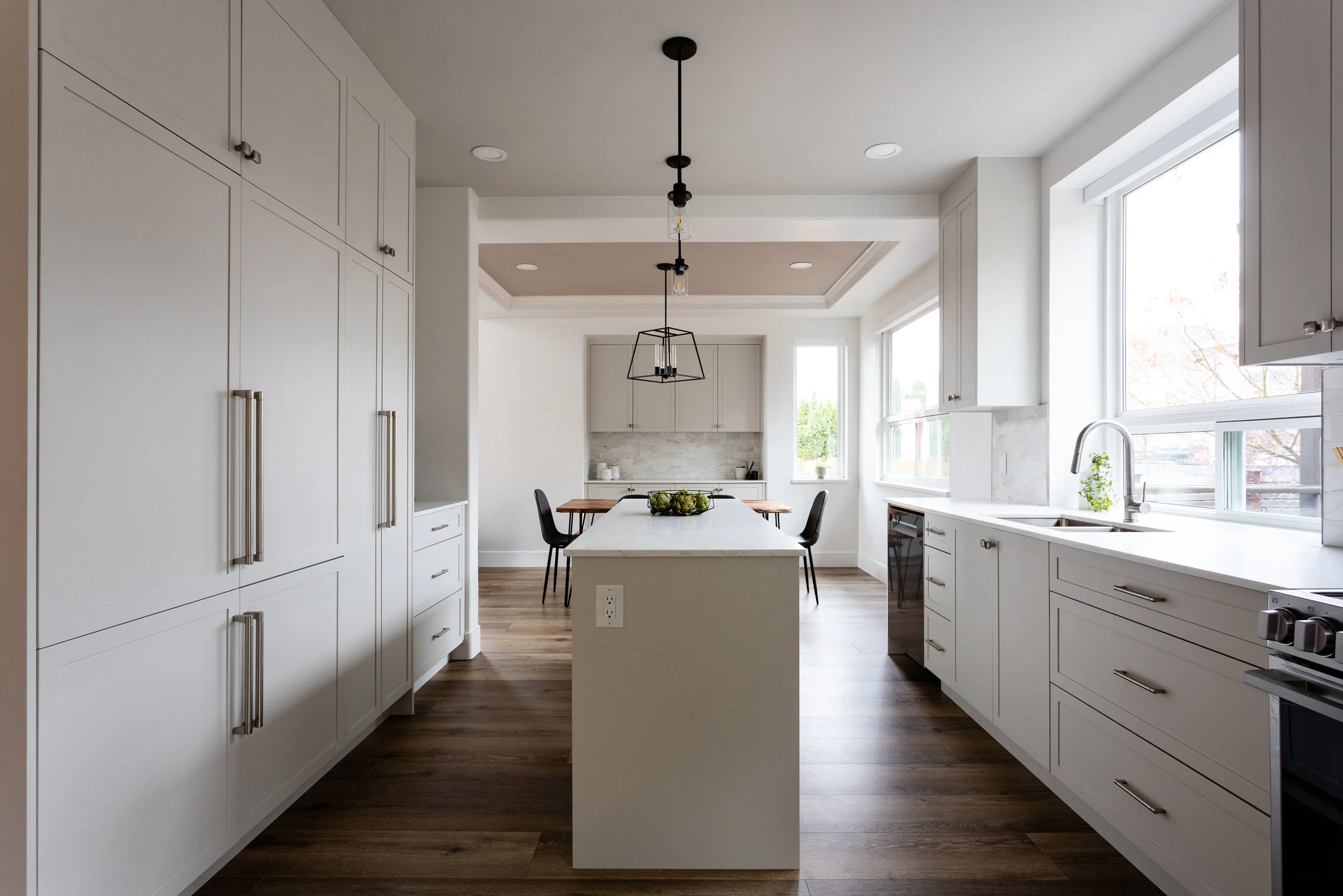 Modern kitchen with white cabinets, island with white countertop, stainless steel appliances, large windows letting in natural light, and wood flooring.