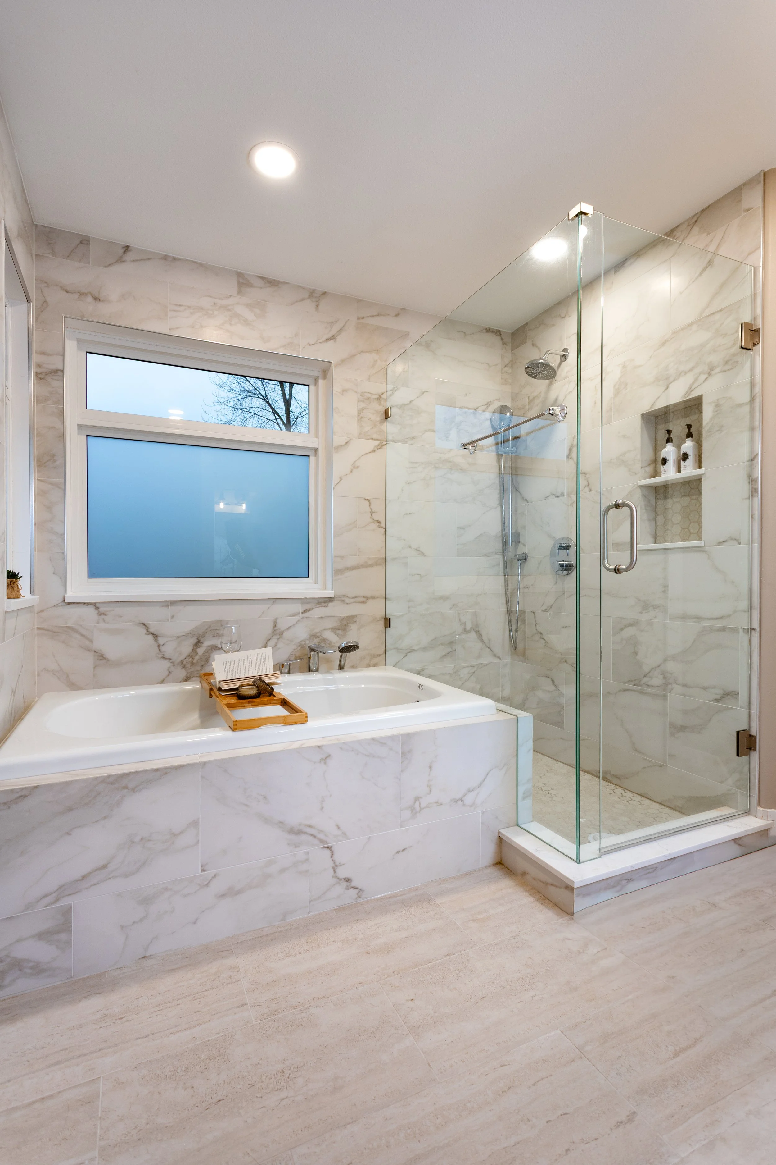 A modern bathroom with a walk-in shower and a bathtub, featuring marble tiles, a frosted window, and a beige-tiled floor.