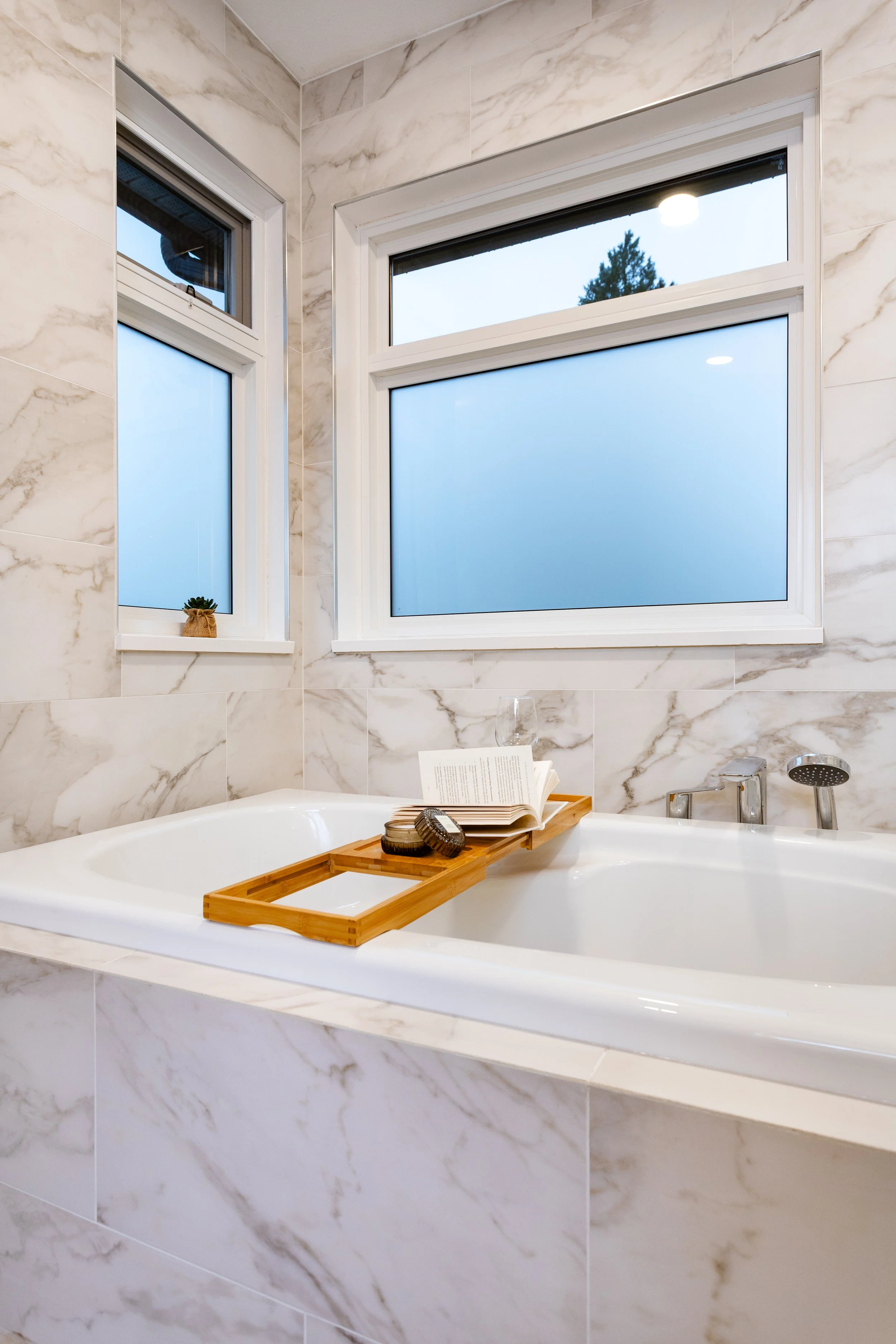 A marble-tiled bathroom with a built-in bathtub, a wooden bath tray holding a book and soap, and two frosted windows allowing natural light to enter.