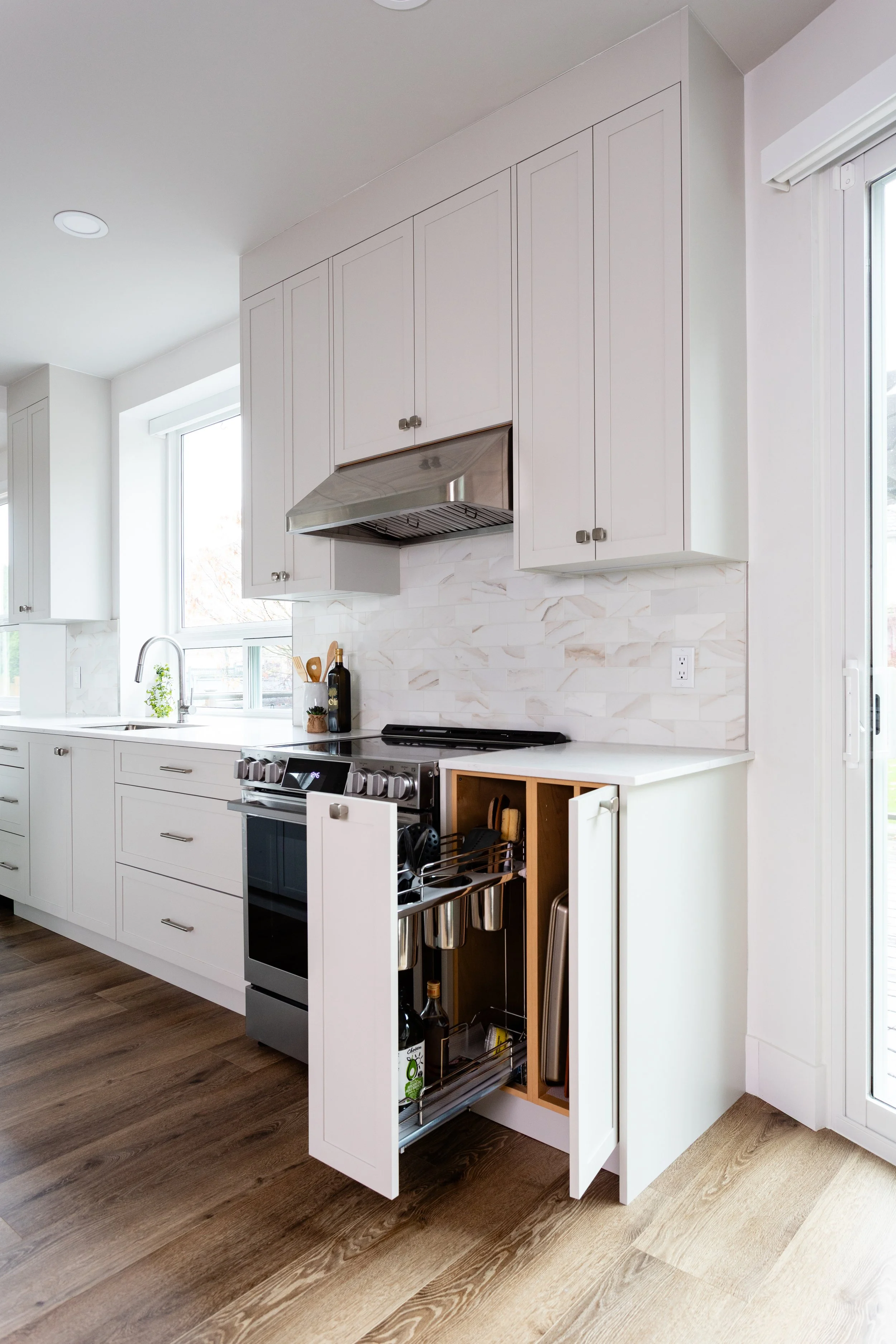 Modern kitchen with white cabinets, stainless steel range hood, and a partially open cabinet revealing hanging utensils and bottles inside. Hardwood floors and a window providing natural light.