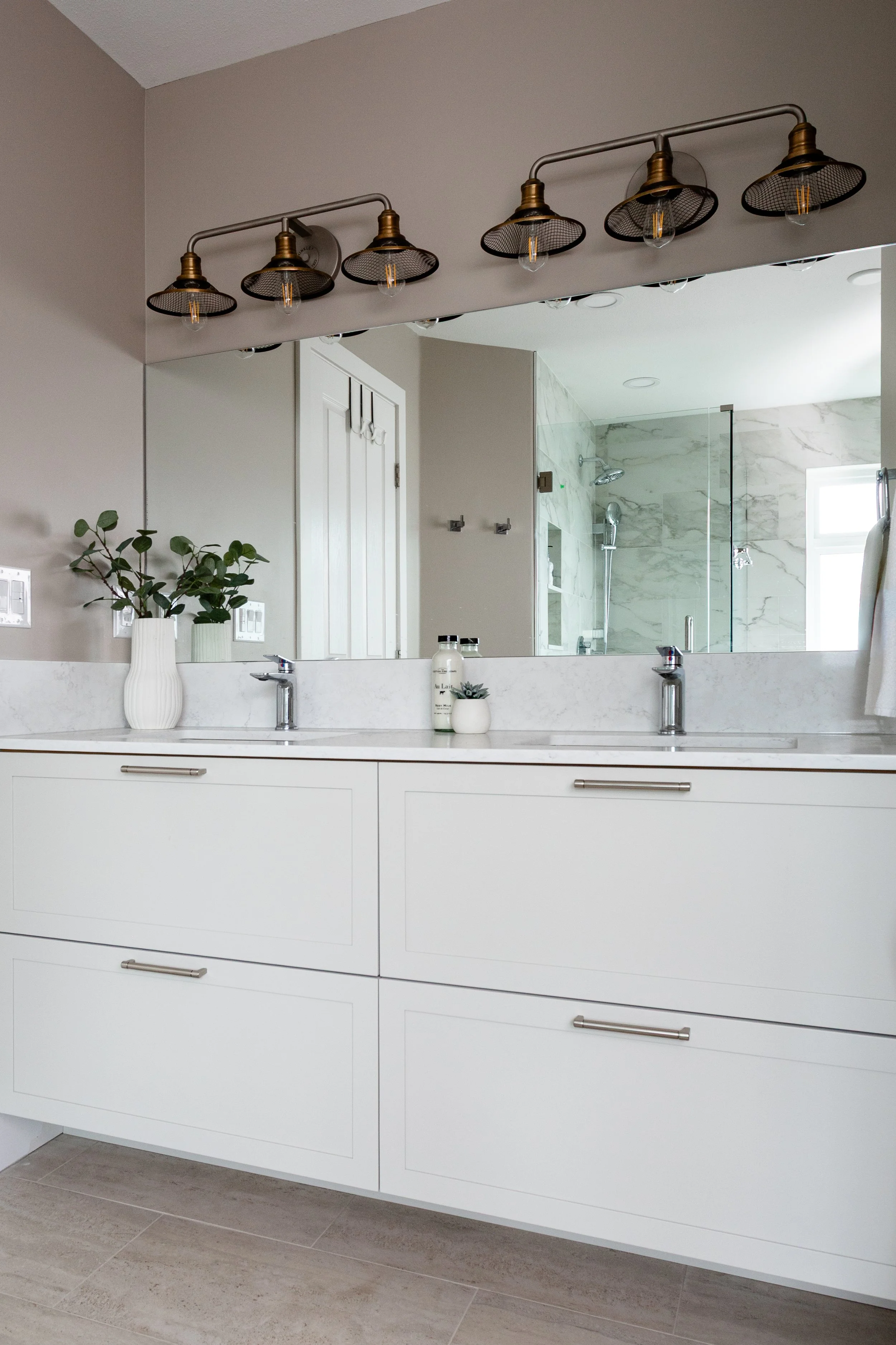 Modern bathroom with white vanity, marble countertop, wall mirror, dual sinks, decorative plants, and overhead light fixtures. Reflection shows walk-in shower with glass door and tiled walls.
