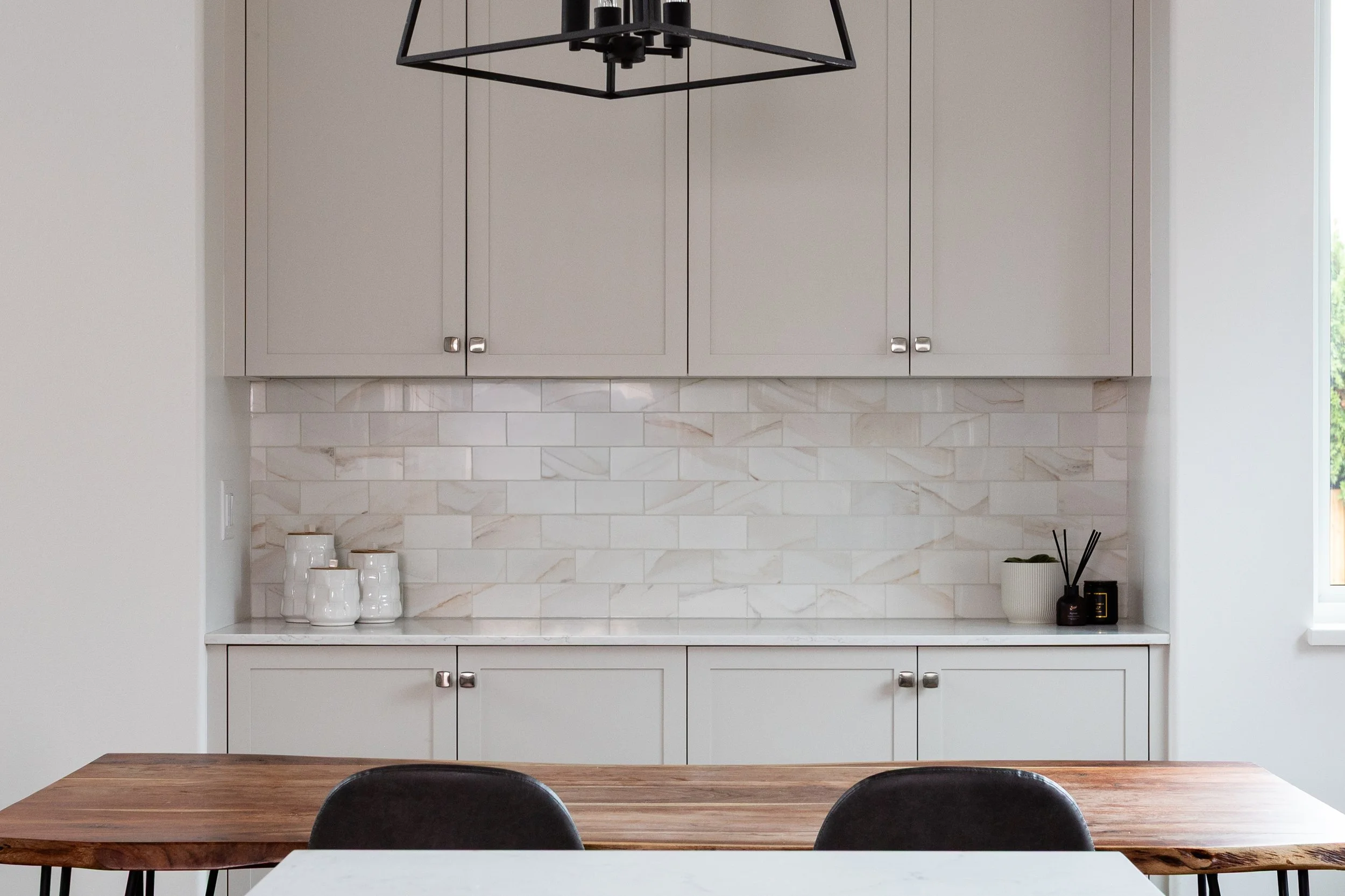 Modern kitchen with gray cabinets, white marble backsplash, wooden dining table, black chairs, and minimalist decor.