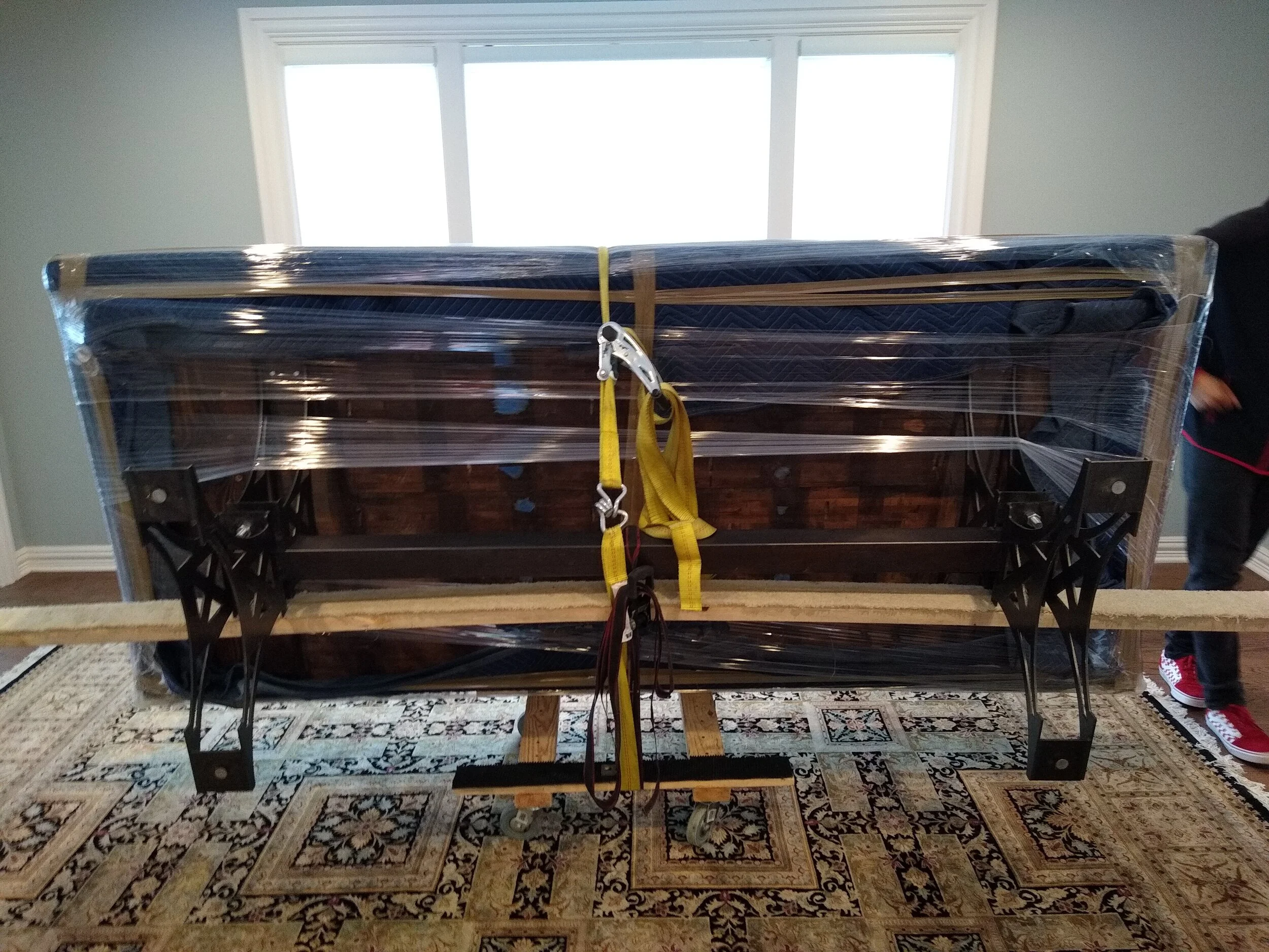 Large wrapped package on a table with black metal stand and wooden supports, tied with yellow straps and plastic wrap, situated in a room with a patterned rug and a window in the background.