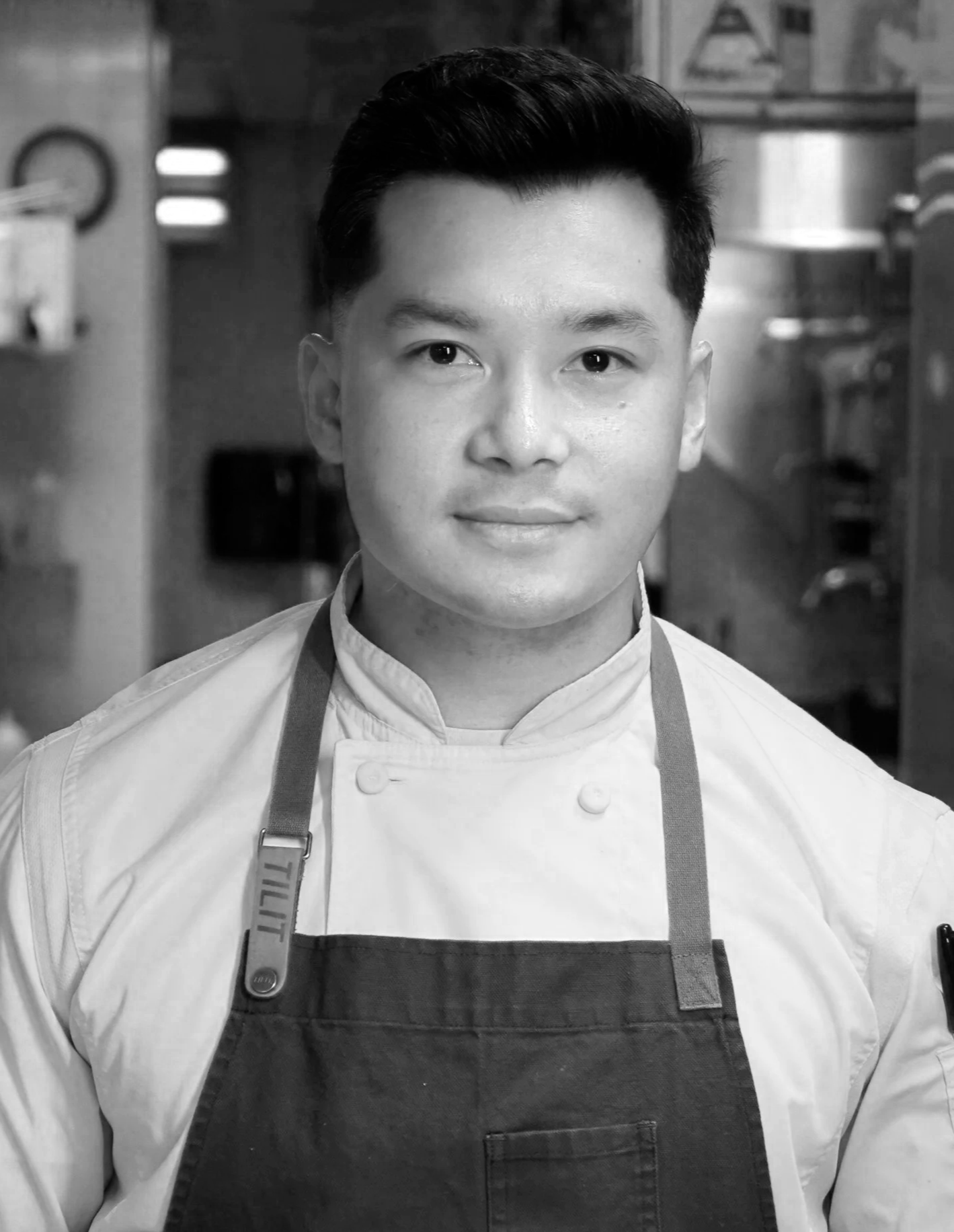 A young male Filipino chef, wearing a white chef's jacket and dark apron, standing in a professional kitchen.