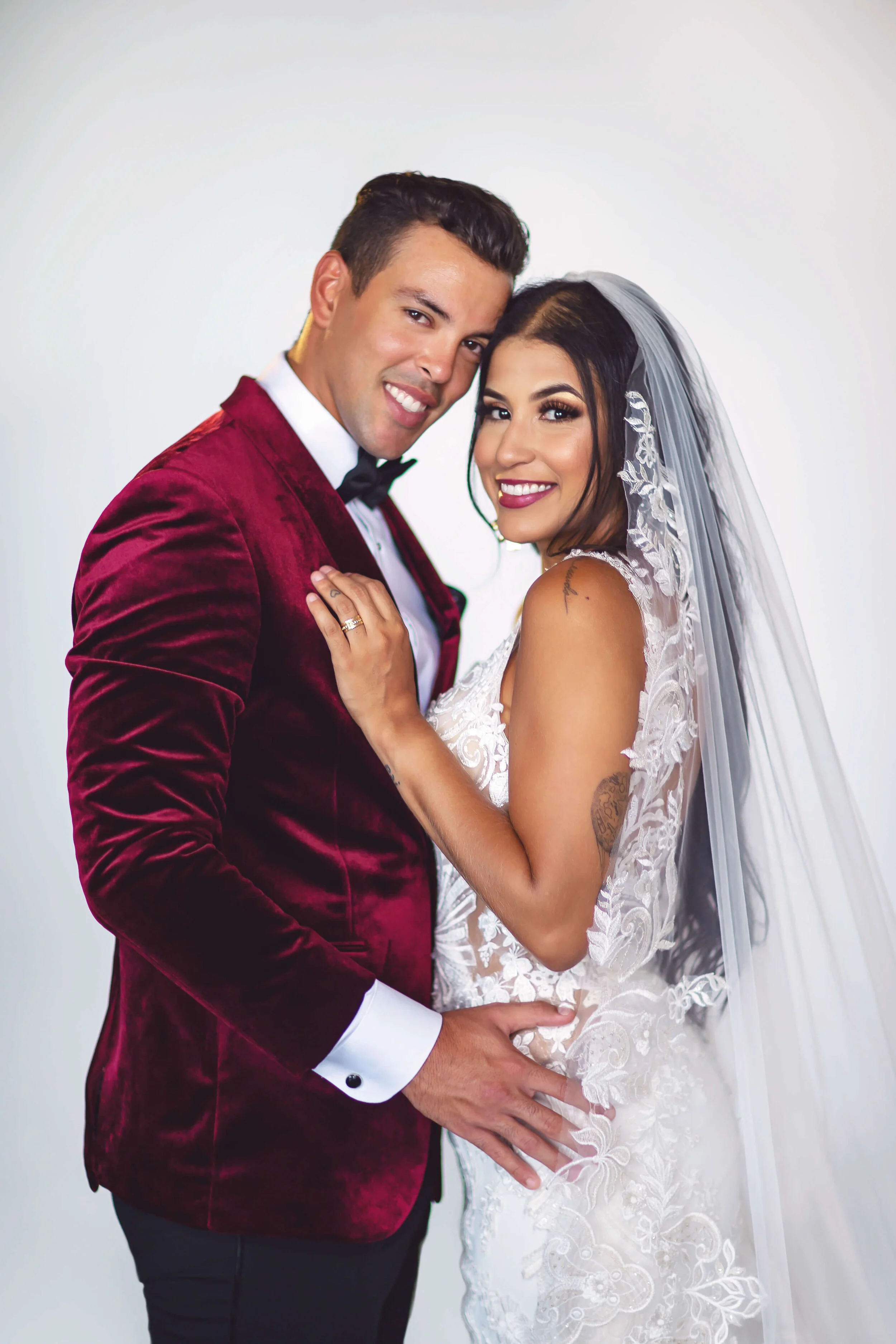 A smiling couple dressed in wedding attire, the man wearing a maroon velvet tuxedo jacket and bowtie, and the woman in a white lace wedding gown with a veil, posing closely together against a plain white background.