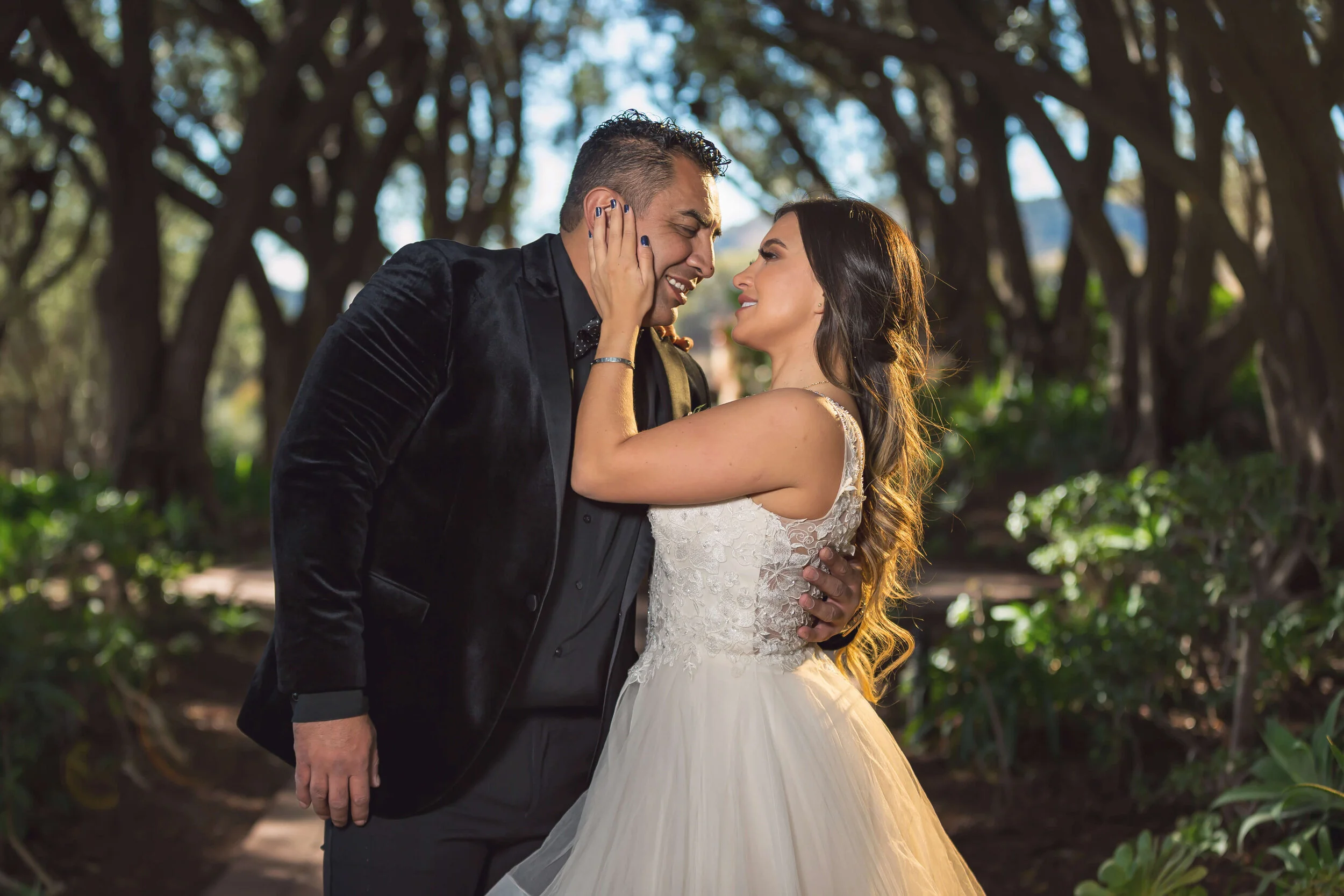 A couple, dressed in wedding attire, sharing an intimate moment in a wooded outdoor setting. The man is wearing a black suit and the woman is in a white wedding dress, with their faces close together and smiling.