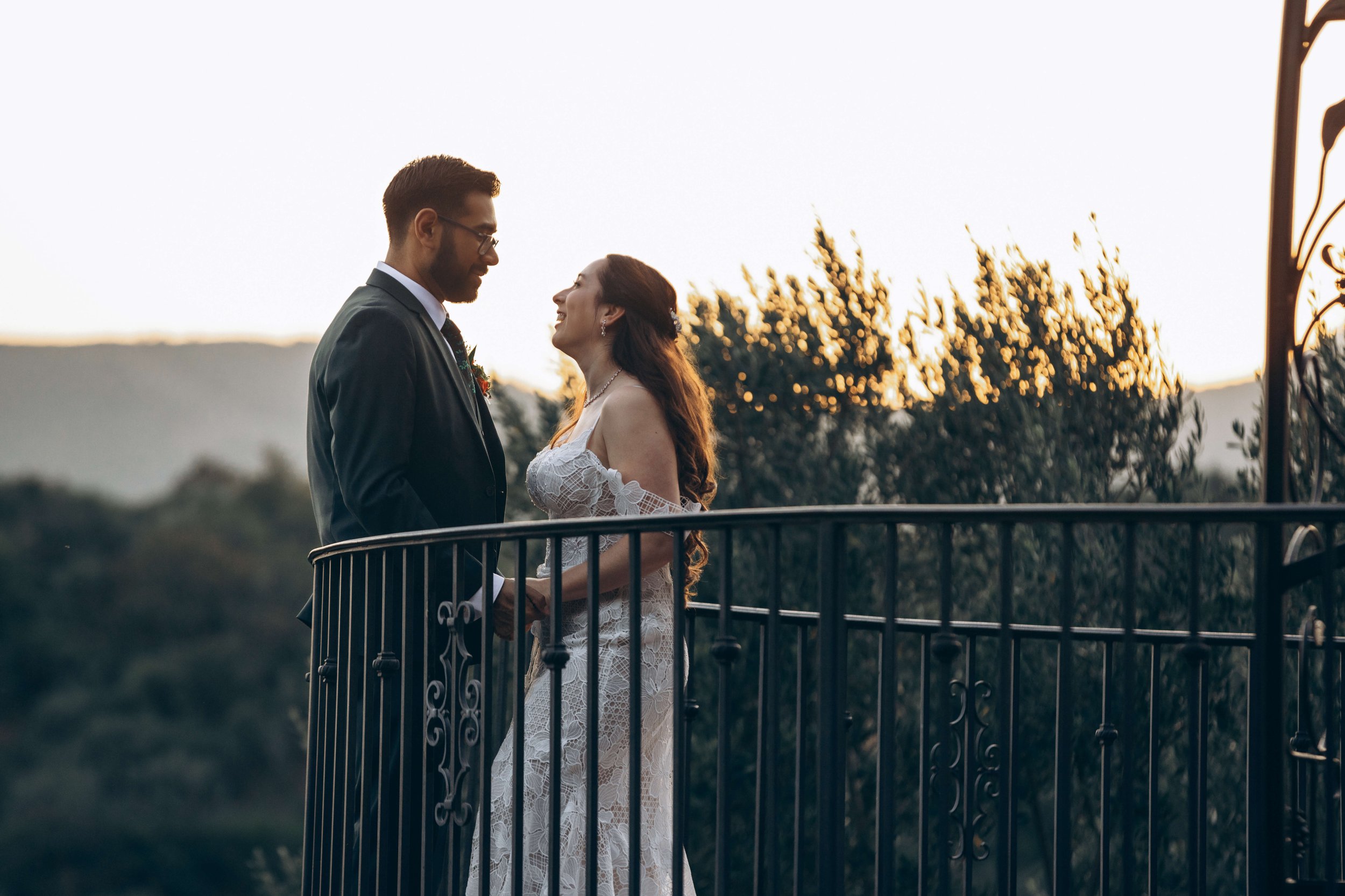 A couple in wedding attire standing on a small balcony, gazing at each other with smiles, during sunset with trees in the background.