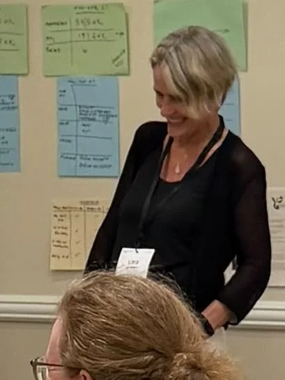 Lisa Bennett with short blonde hair wearing a black top and lanyard, standing and smiling in front of a wall with colorful notes or papers.