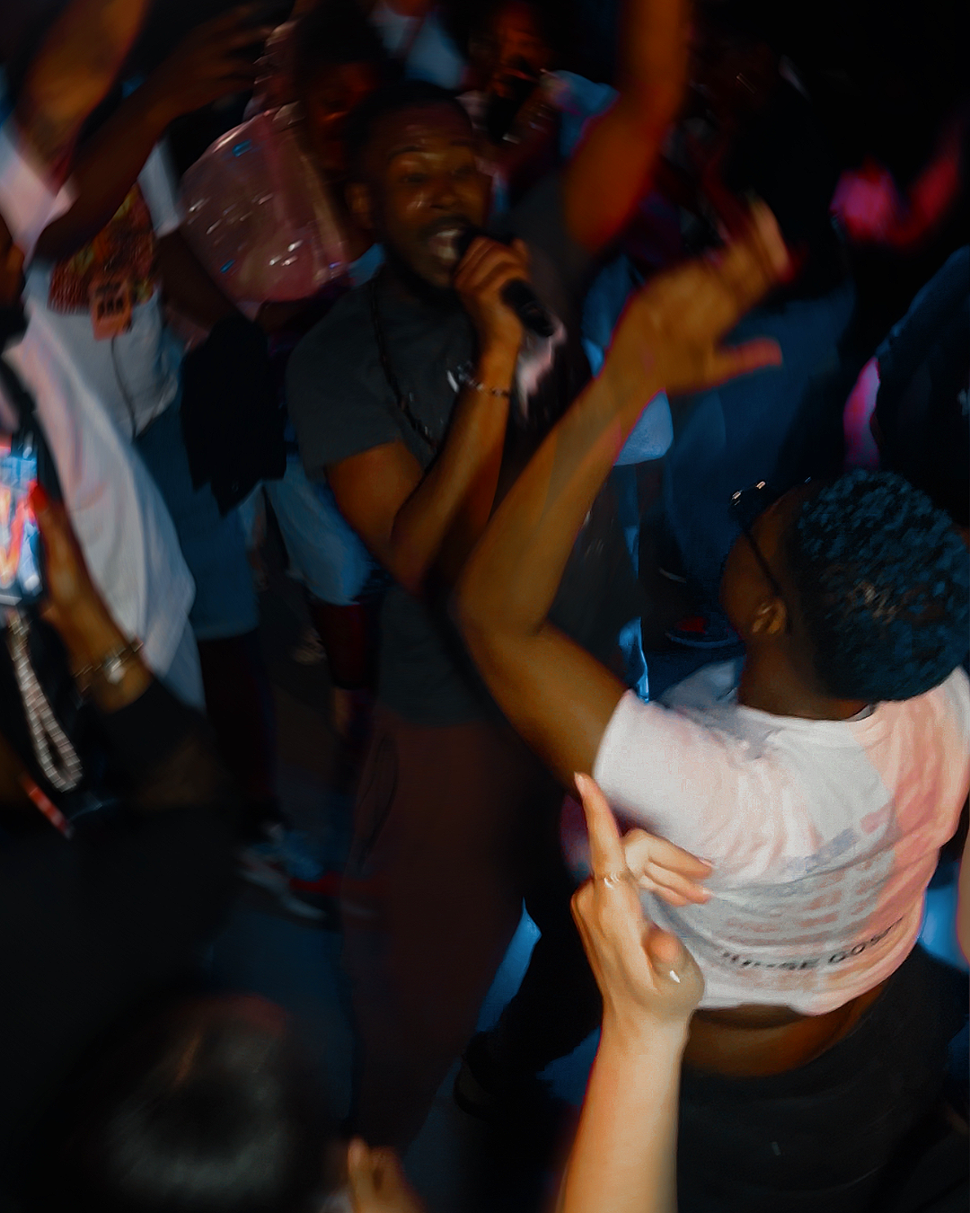 A photograph of lots of people on a dance floor at a rave, centred around one man with his arm in the air and the other hand holding a microphone