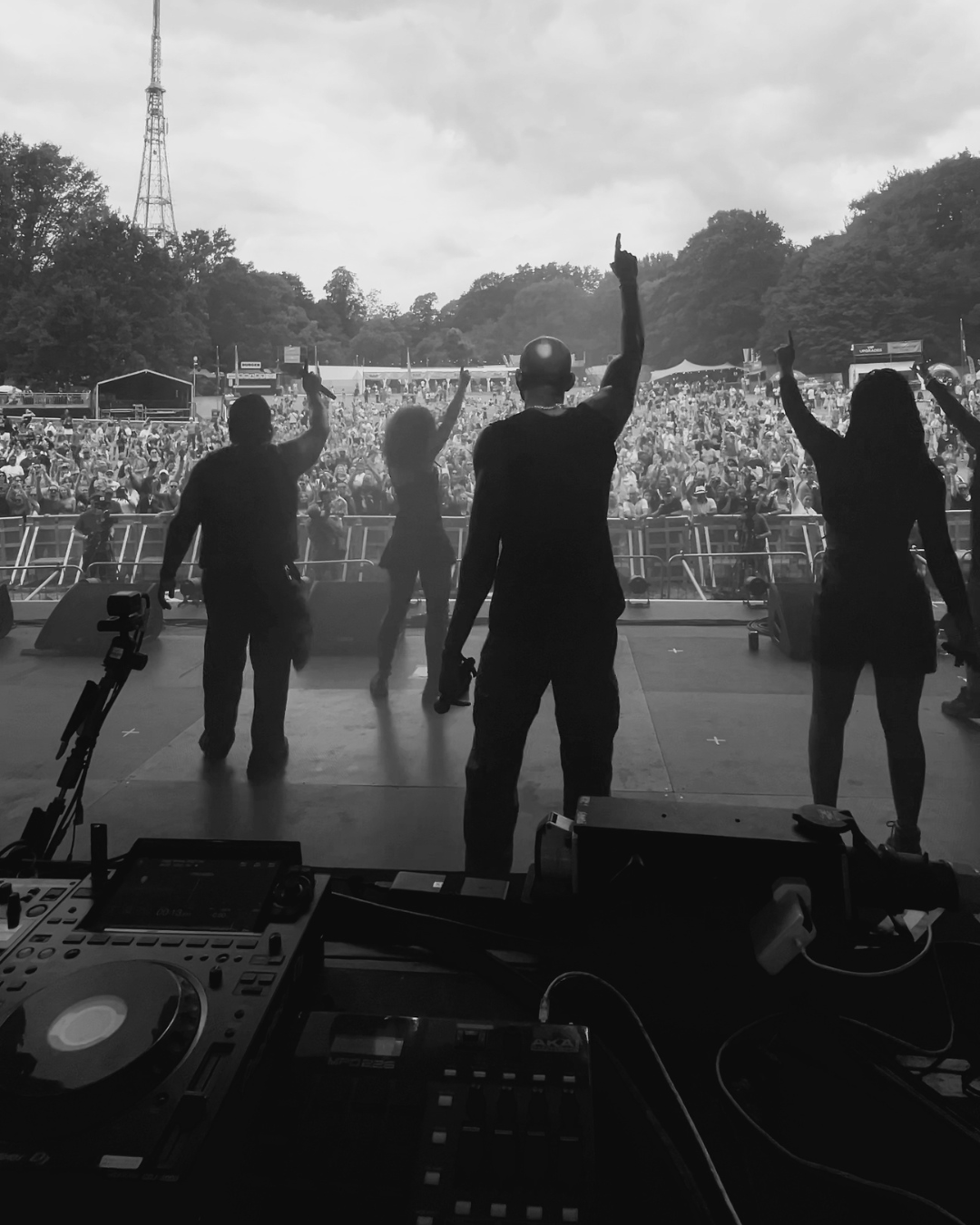 Photograph from behind the DJ Booth looking out onto a large crowd at a music festival. In the foreground are DJ decks and between them and the crowd are several singers all with one arm in the air holding up one finger to unite the crowd infront