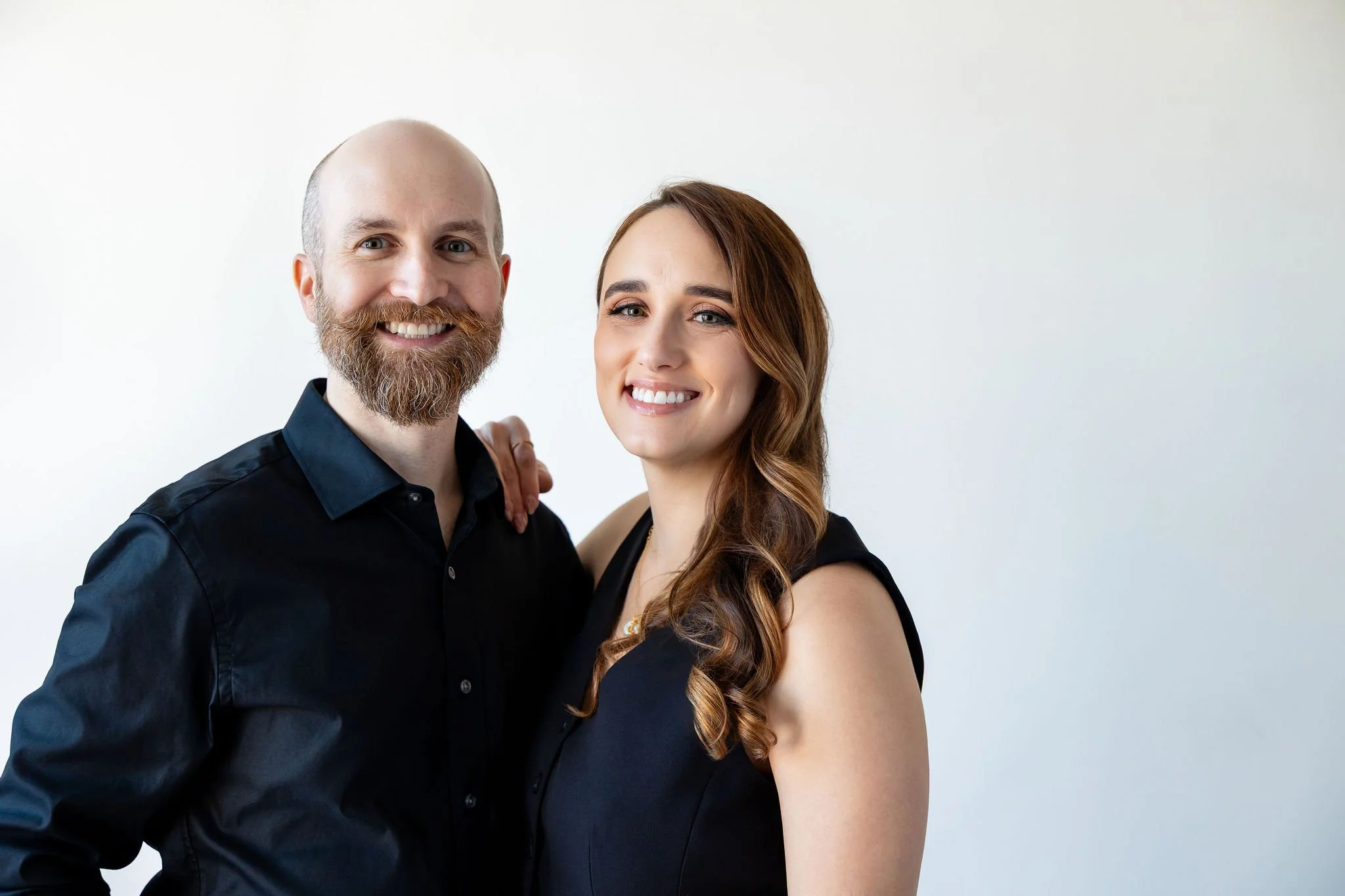 A smiling man with a beard and a woman with long hair, both dressed in black, posing together against a plain white background.