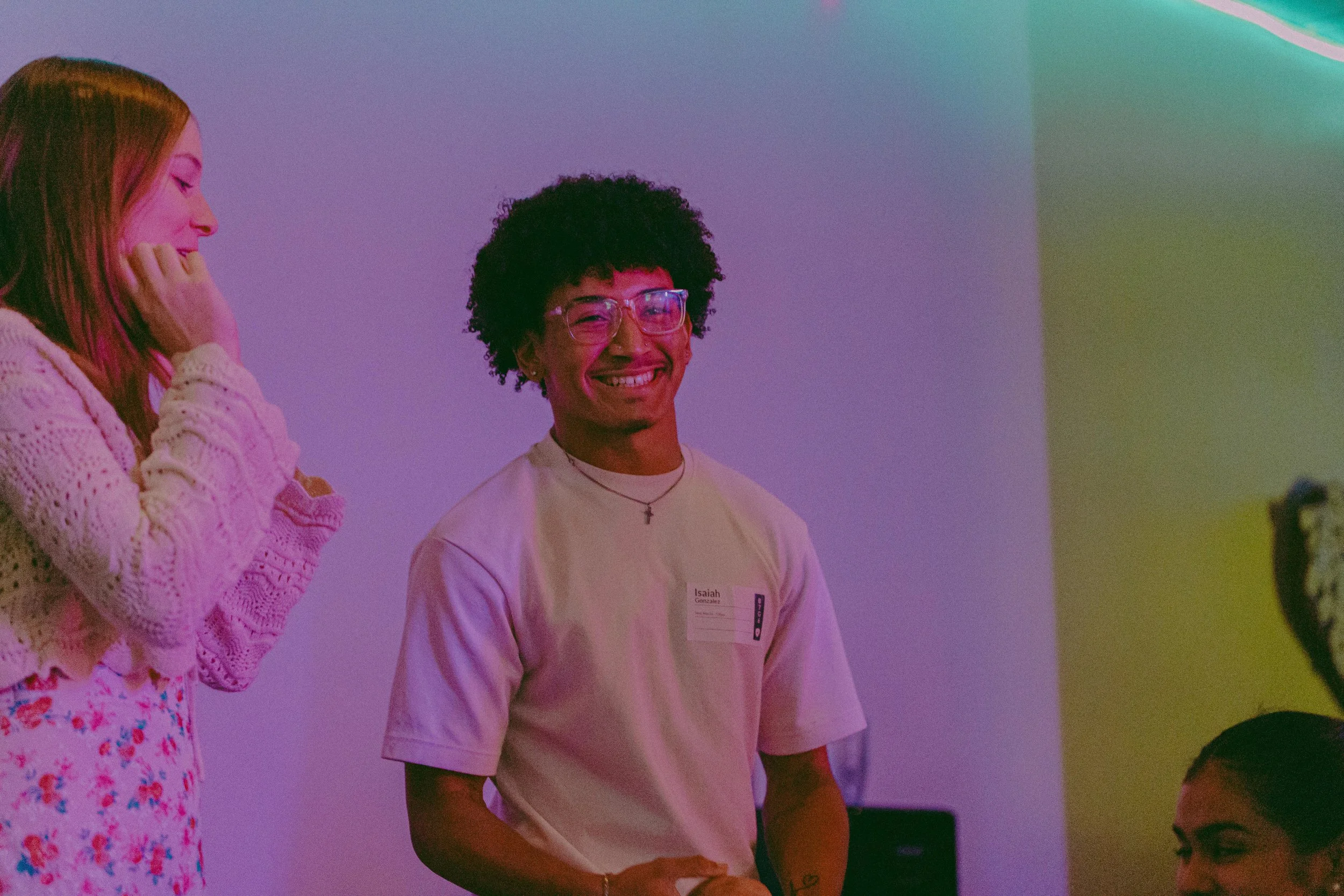 Smiling young man with curly black hair, glasses, and a beige t-shirt standing next to women, one with red hair and one with dark hair, in a room with colorful lighting.