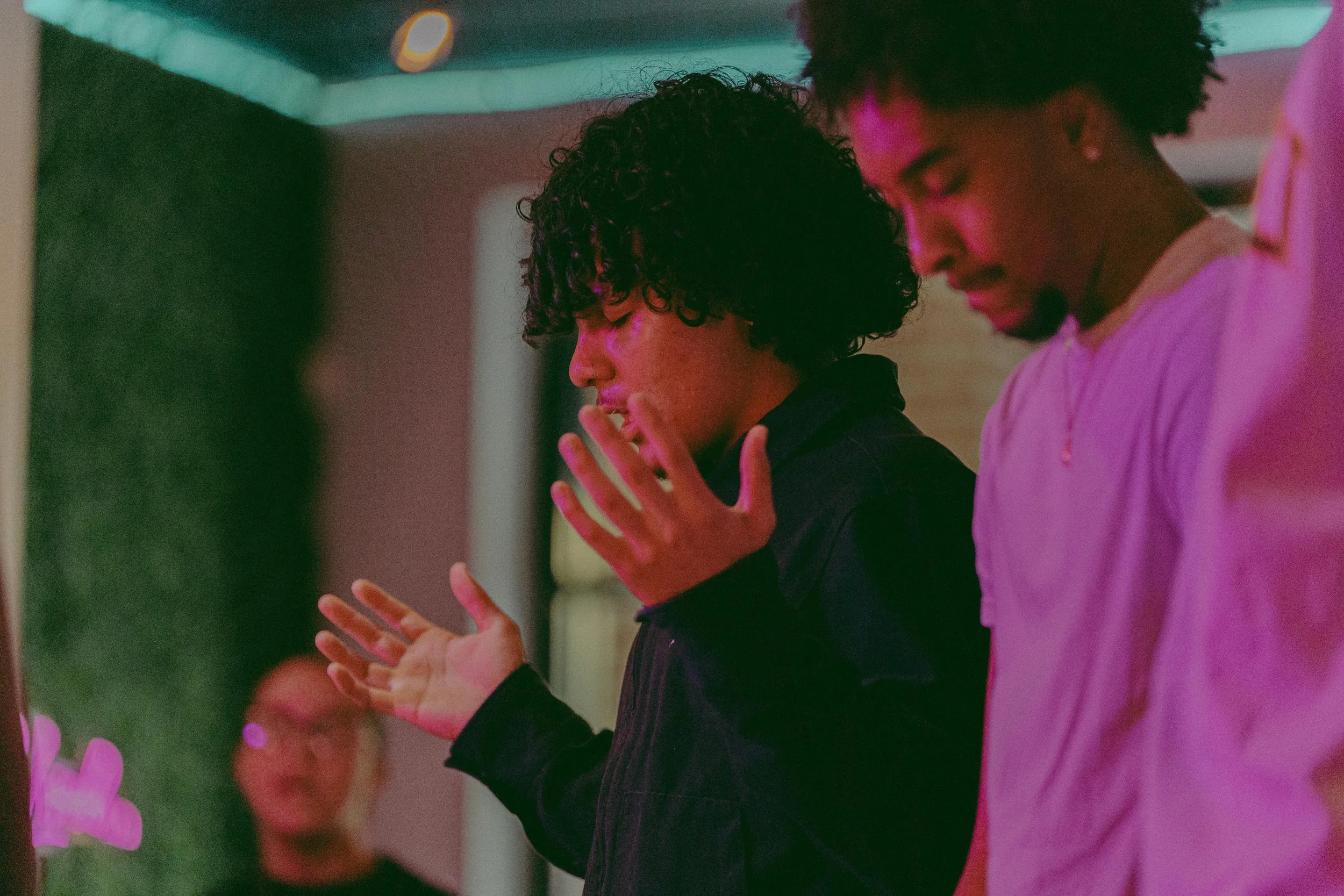 Two young men with curly hair praying with their eyes closed and hands raised in an indoor setting with pink and purple lighting.