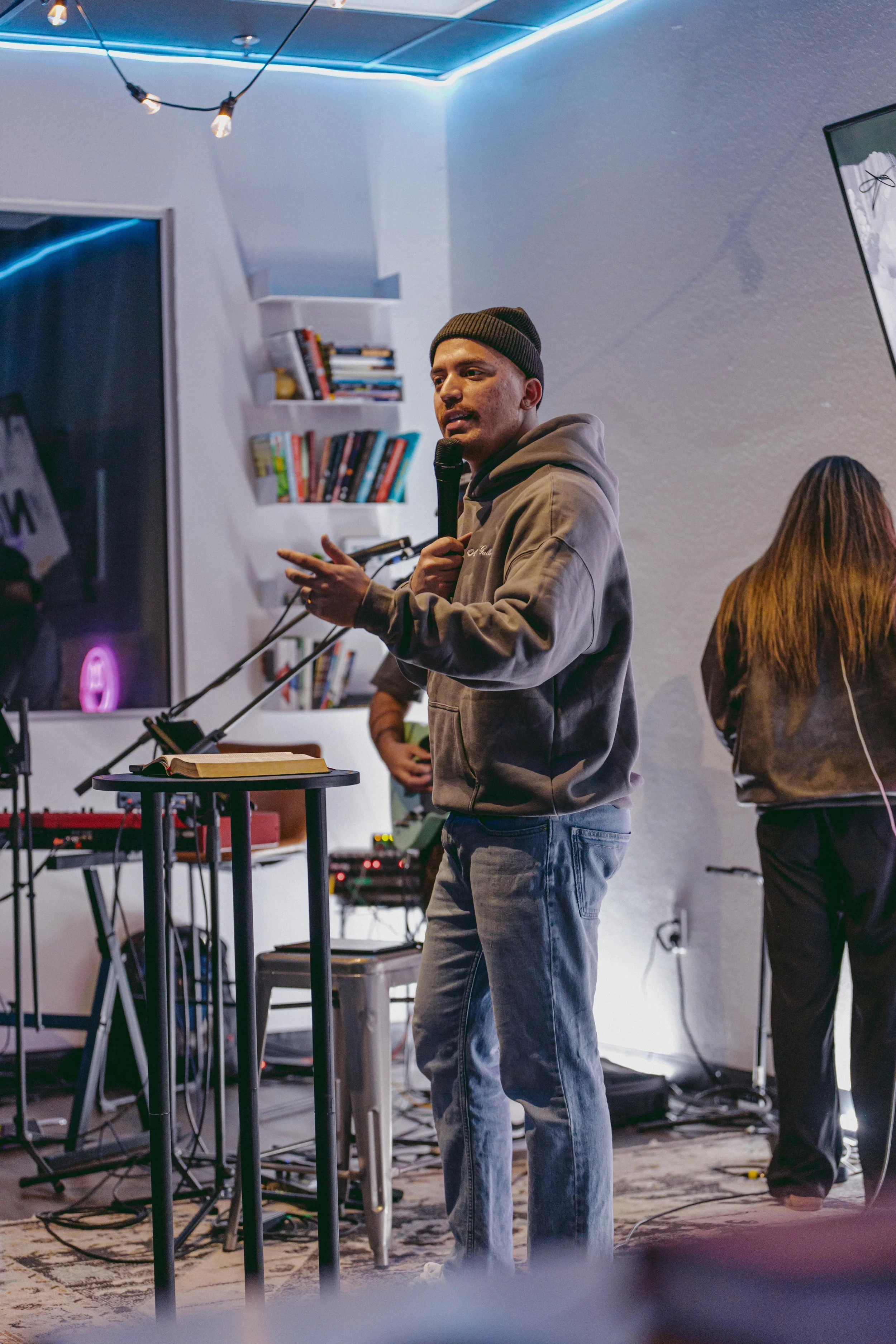 A man wearing a beanie and a hoodie speaking into a microphone during a presentation or event in a cozy room with blue lighting, keyboard, and bookshelves.