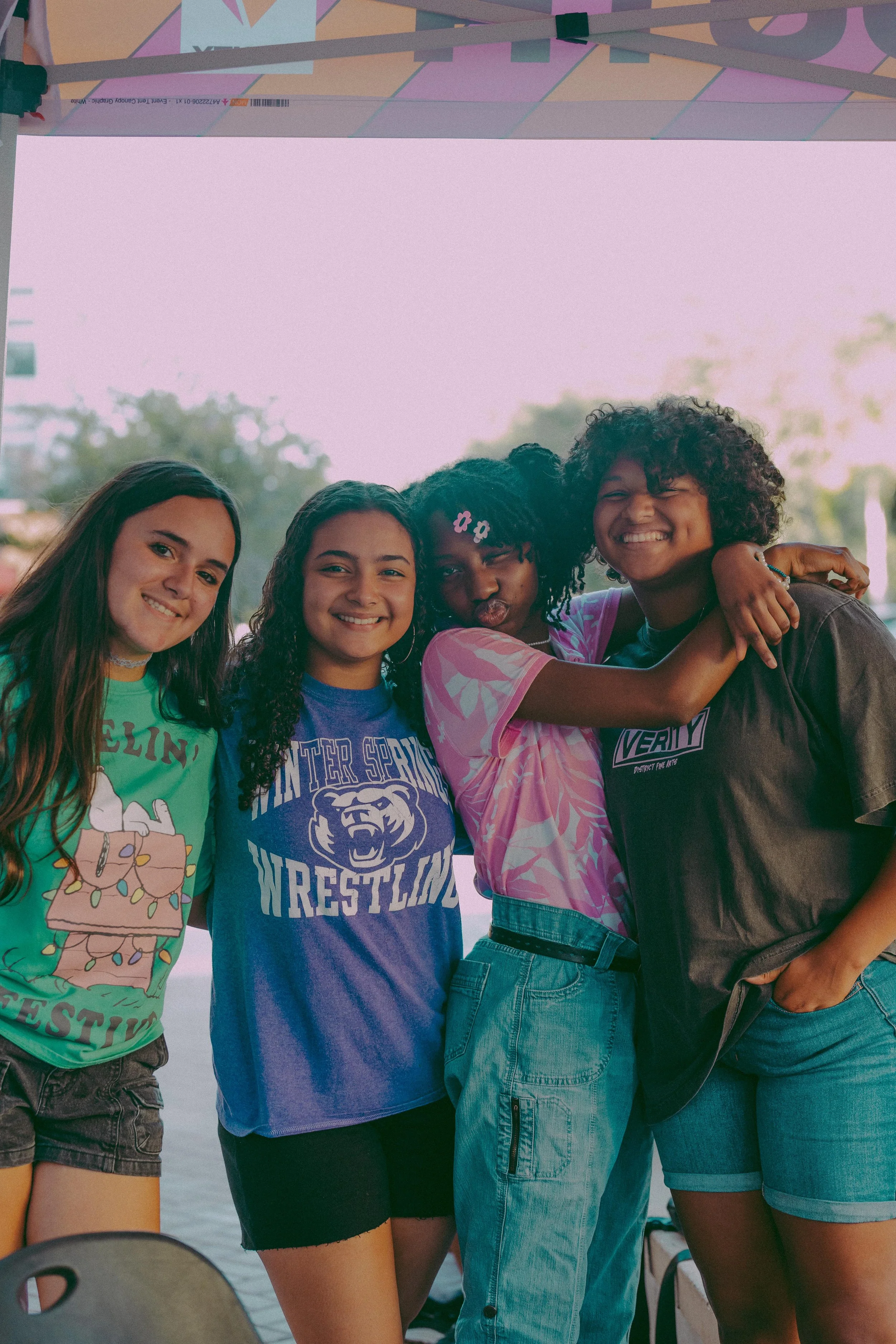 Four girls standing together outdoors, smiling, with one hugging another. They are wearing colorful casual clothing, including a tie-dye shirt, a wrestling T-shirt, and a festive graphic T-shirt. The background shows trees and a cloudy sky.