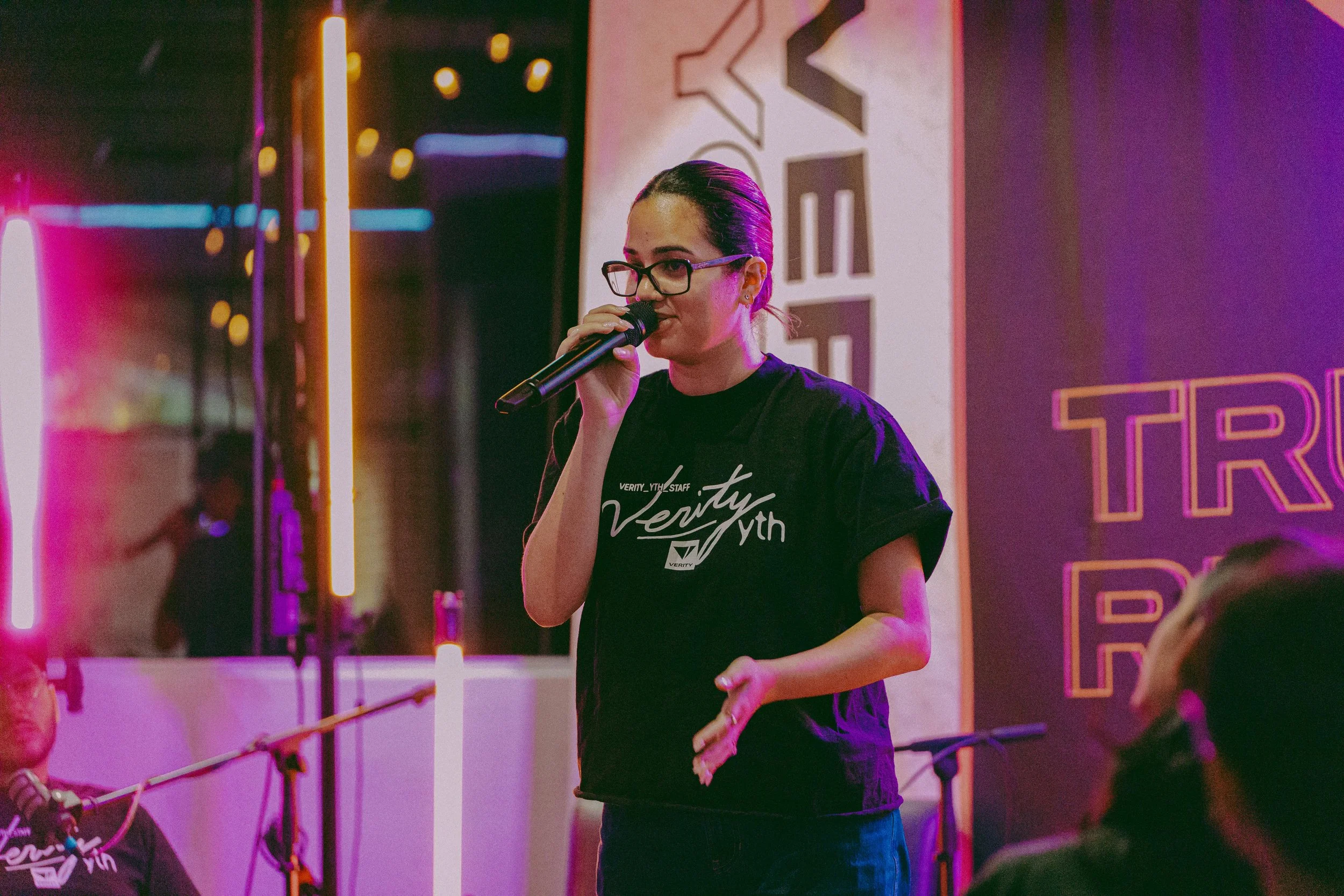 A woman with glasses and dark hair tied back, wearing a black T-shirt, speaking or singing into a microphone during an event with purple and pink neon lighting.
