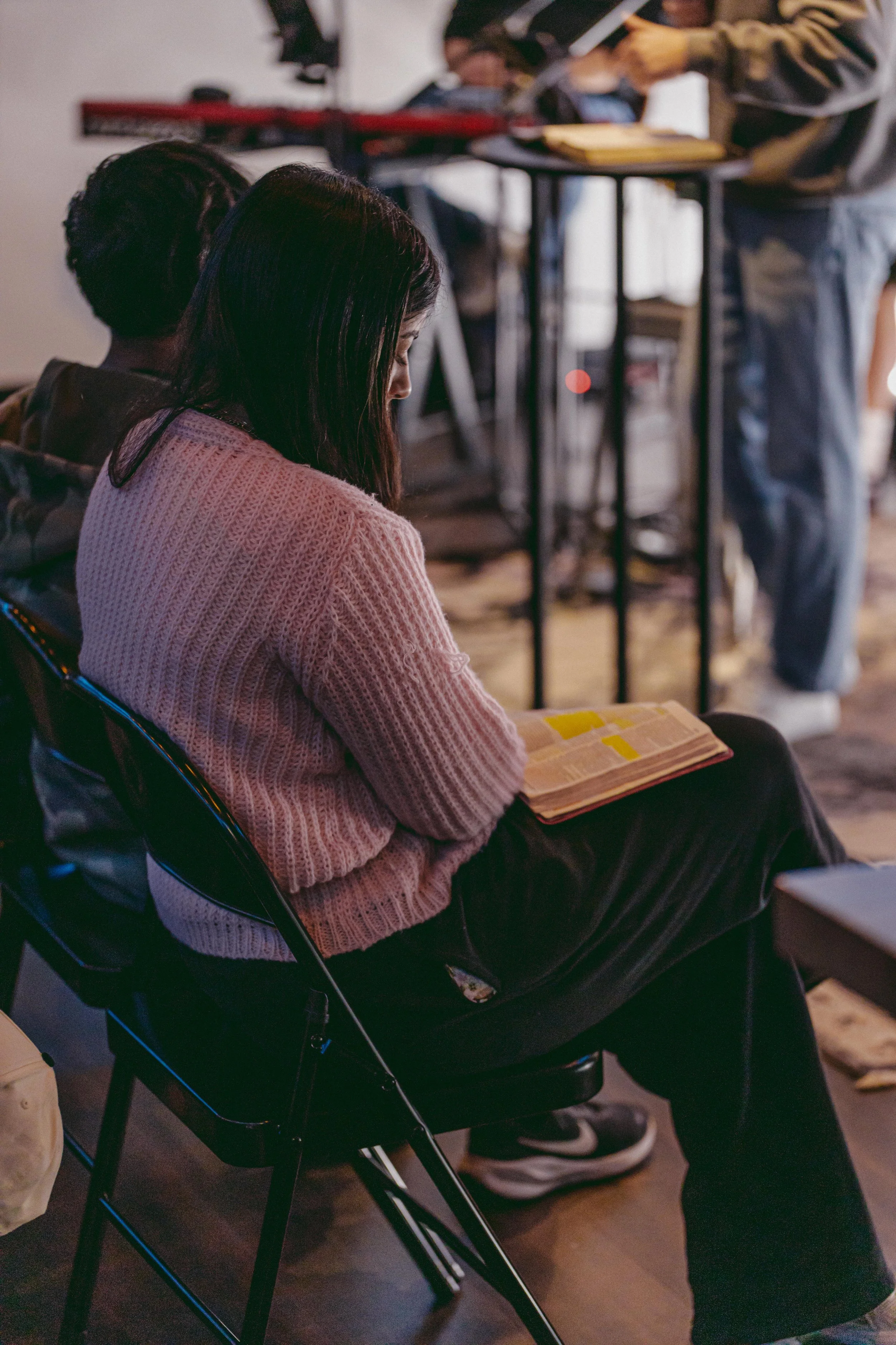 A woman with long dark hair wearing a pink sweater sitting on a black chair, reading a book with yellow highlights in a room with other people and a person standing in the background.
