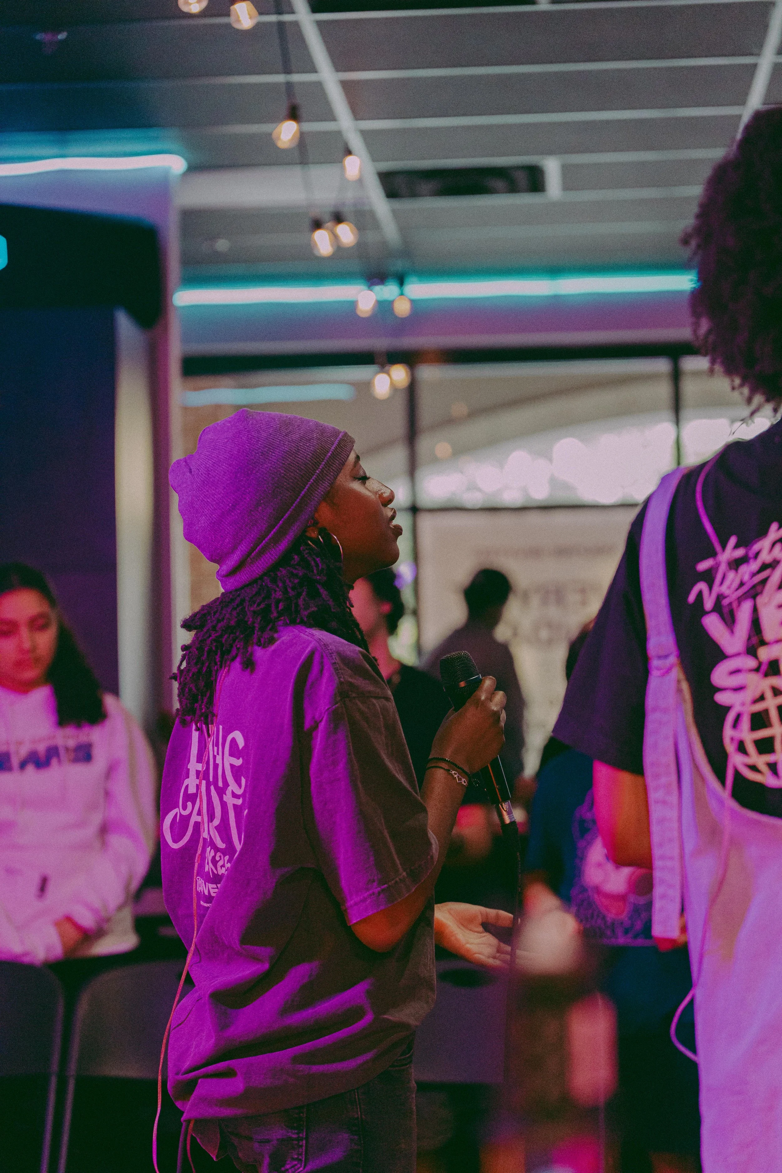 A young woman with dreadlocks, wearing a gray beanie and a gray T-shirt, holding a microphone, standing in a bustling indoor space with neon lights and other people in the background.