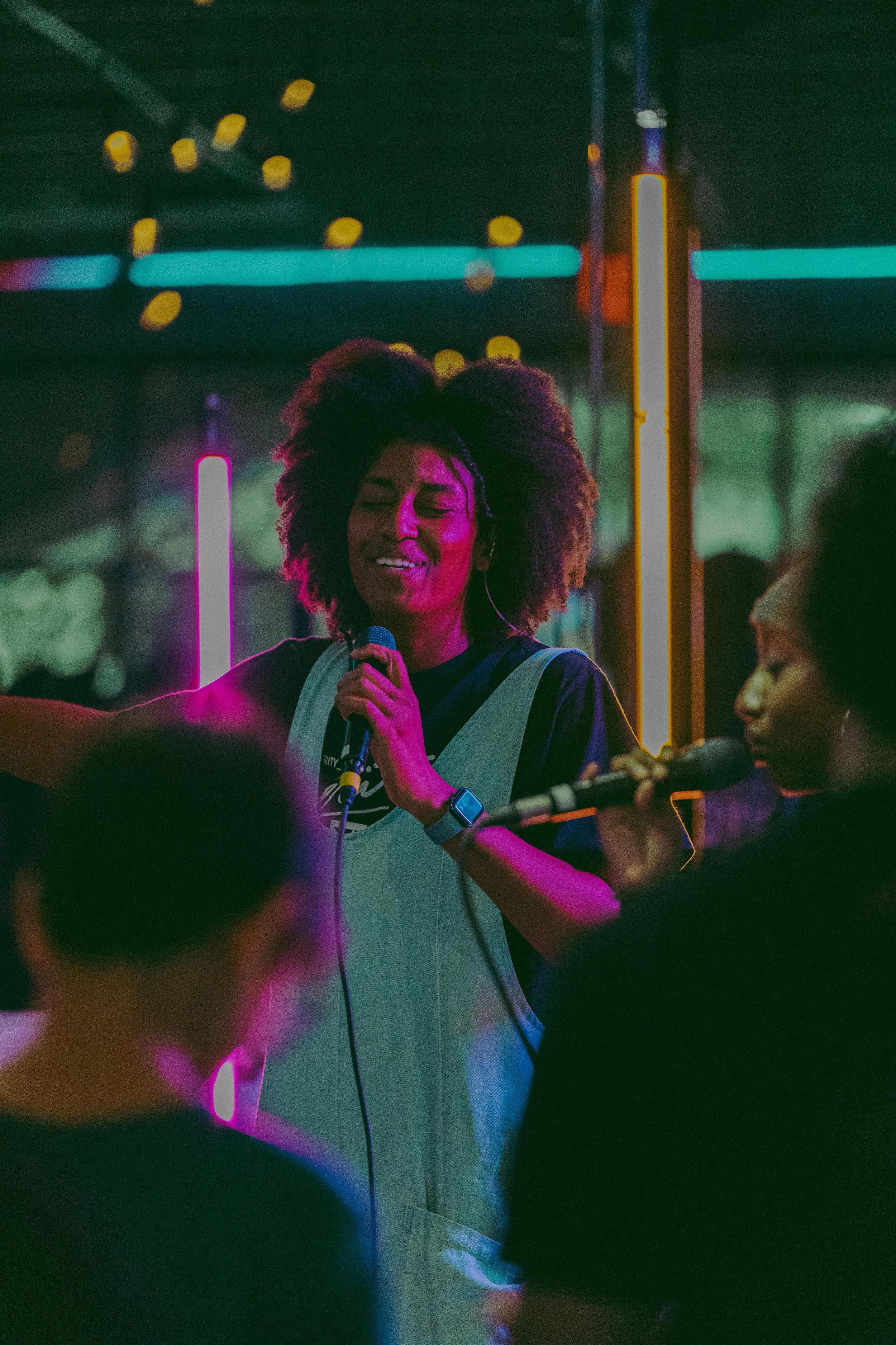 A woman with curly hair singing into a microphone during a live performance at a venue with colorful neon lights.