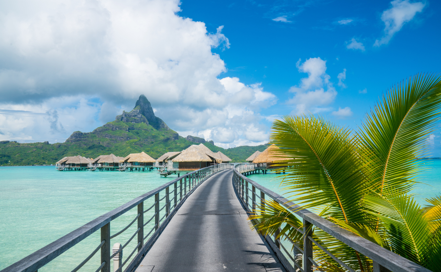 Overwater bungalows connected by a boardwalk with a lush mountain rising behind them.