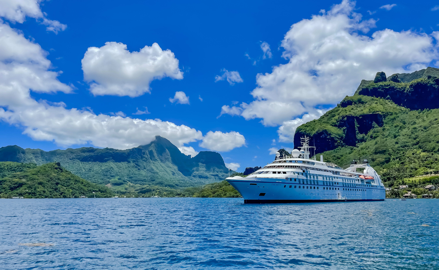 A small luxury cruise ship sailing through calm blue waters with lush green mountains in the background in French Polynesia.