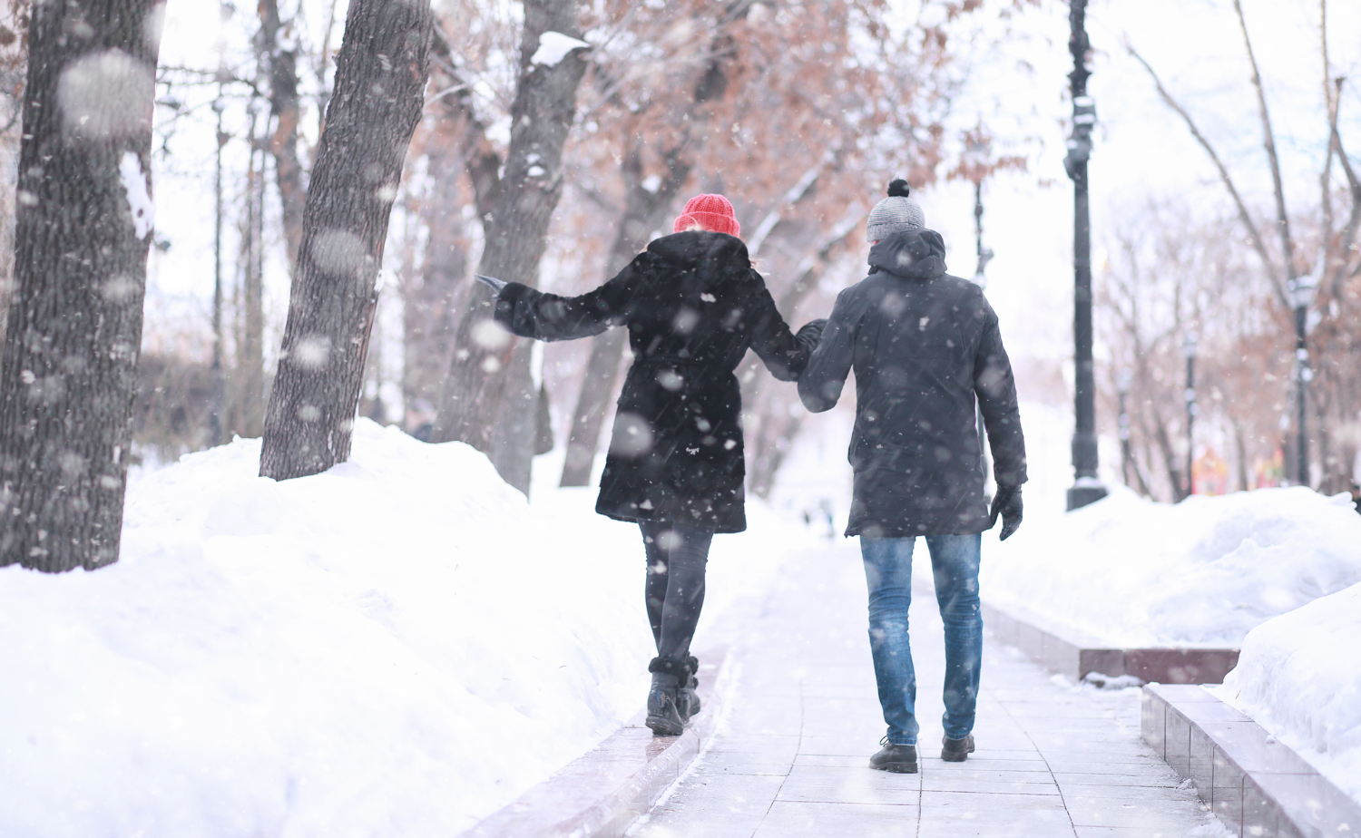 A couple walking hand in hand along a snowy path, surrounded by falling snow and winter trees