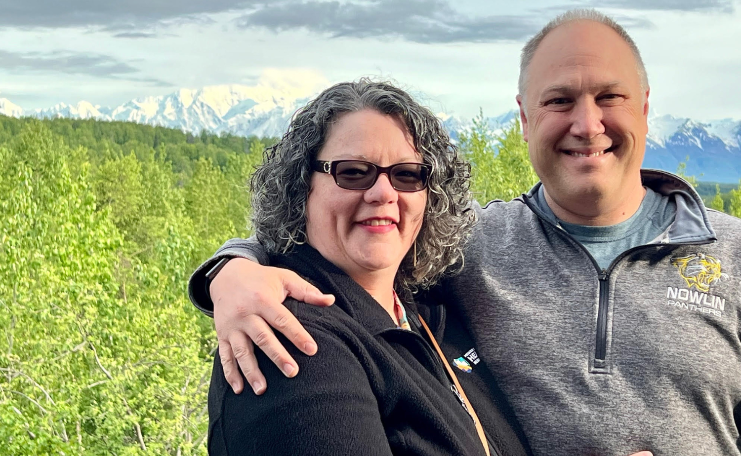 Brian and Jenny smiling in front of lush green trees and snow-capped peaks during their Alaska adventure
