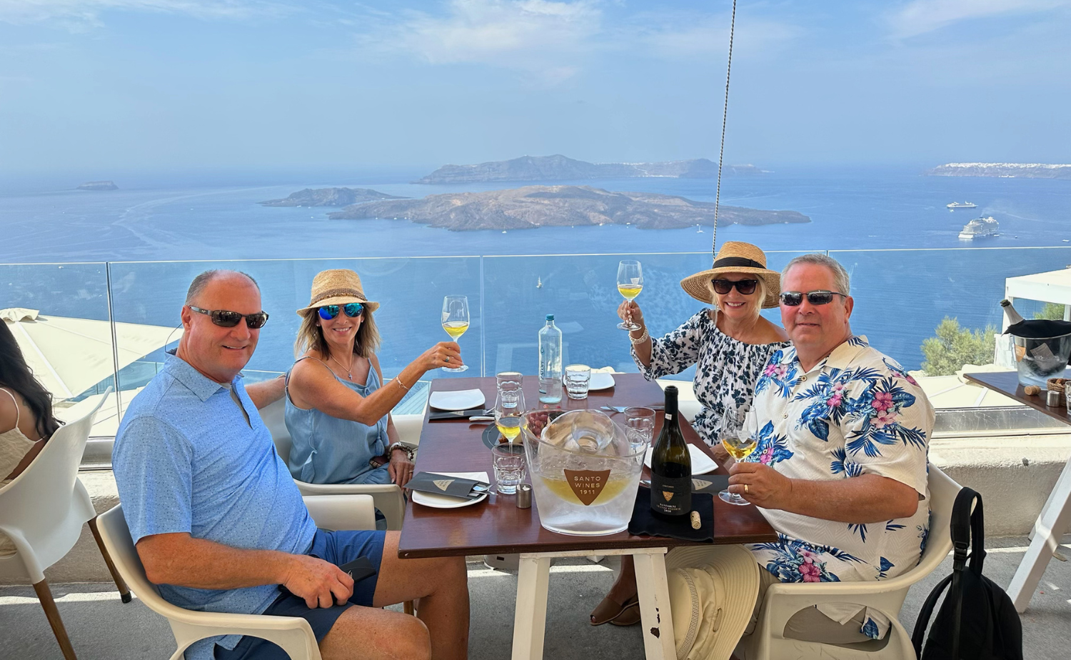 Four friends toasting glasses of white wine at an outdoor table with a stunning view of the Santorini caldera.