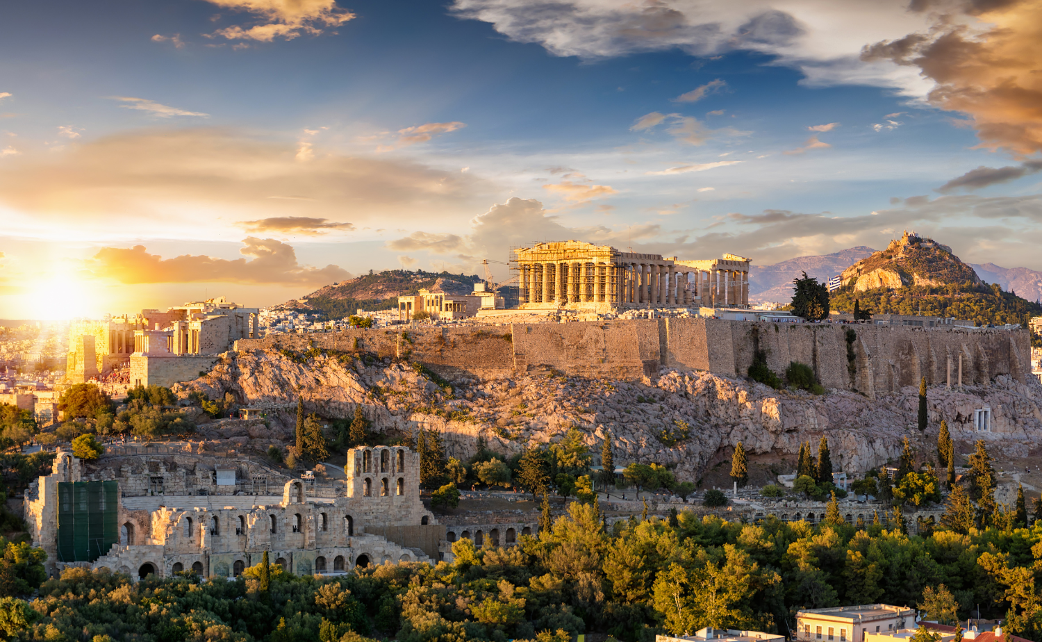 View of the Acropolis of Athens at sunset, with the Parthenon and surrounding ancient ruins glowing in golden light.