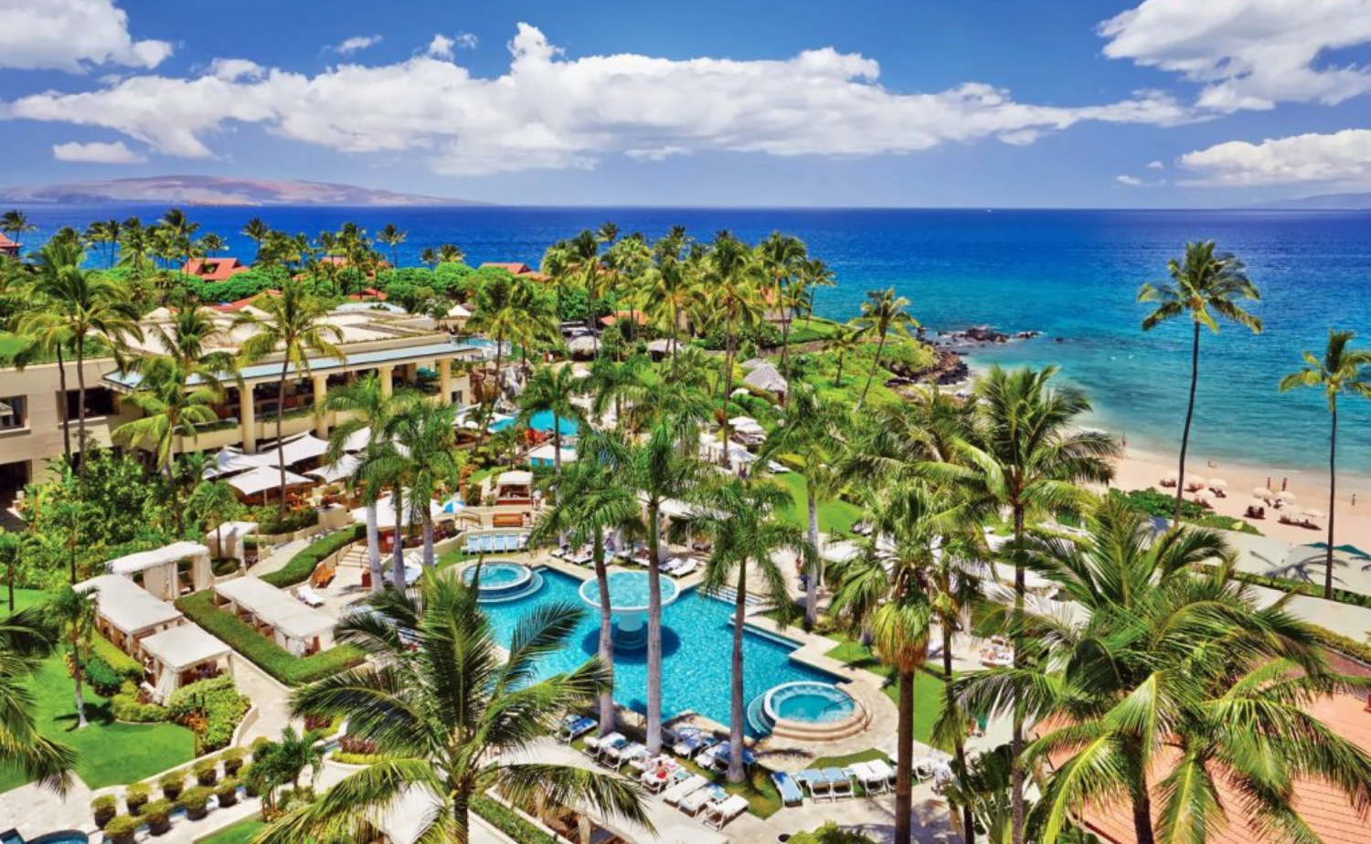 Aerial view of a luxury beachfront resort in Hawaii with palm trees, pools, and the ocean beyond.