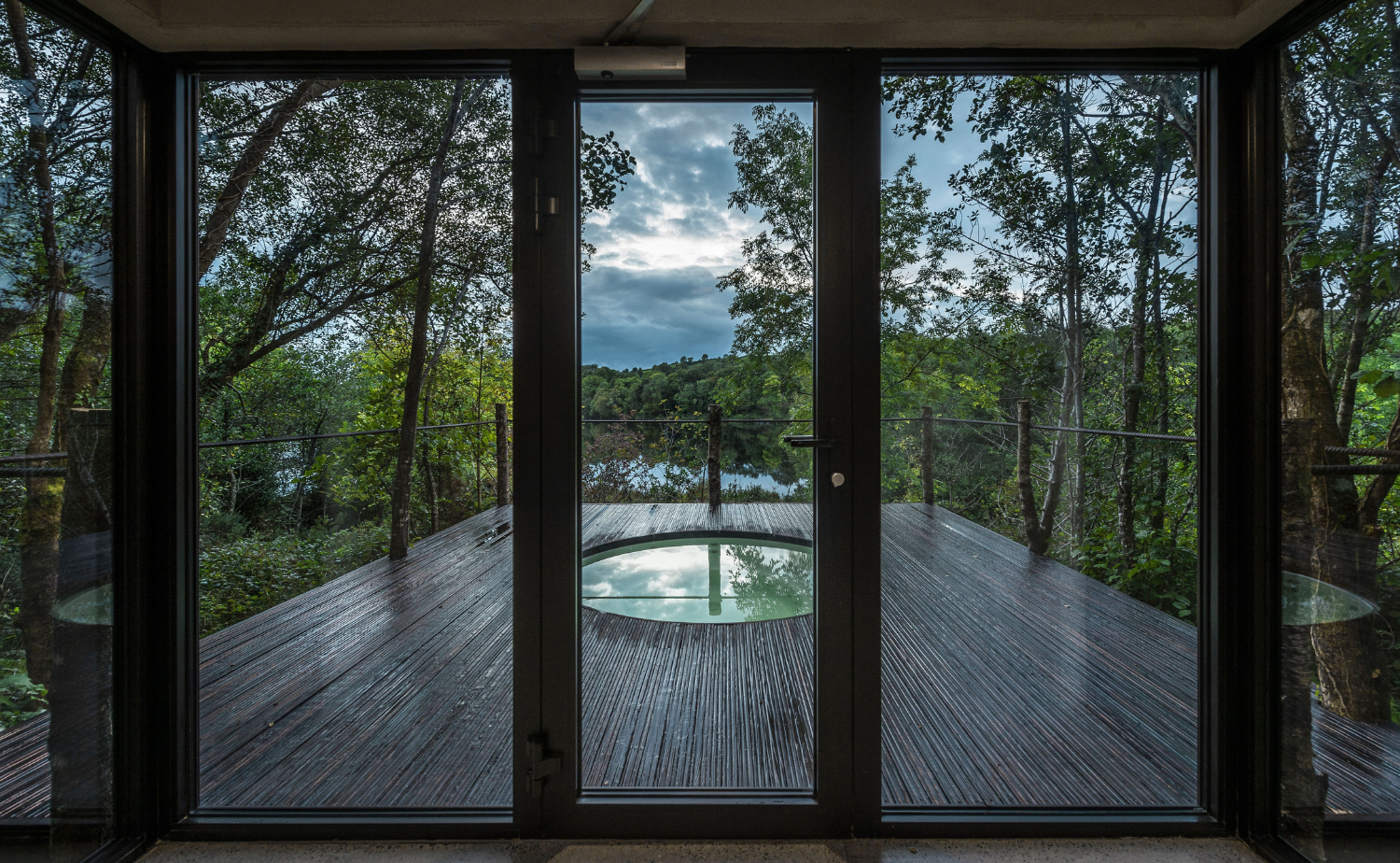 View from inside a Finn Lough spa suite looking out to a private outdoor hot tub deck surrounded by trees and overlooking the water.