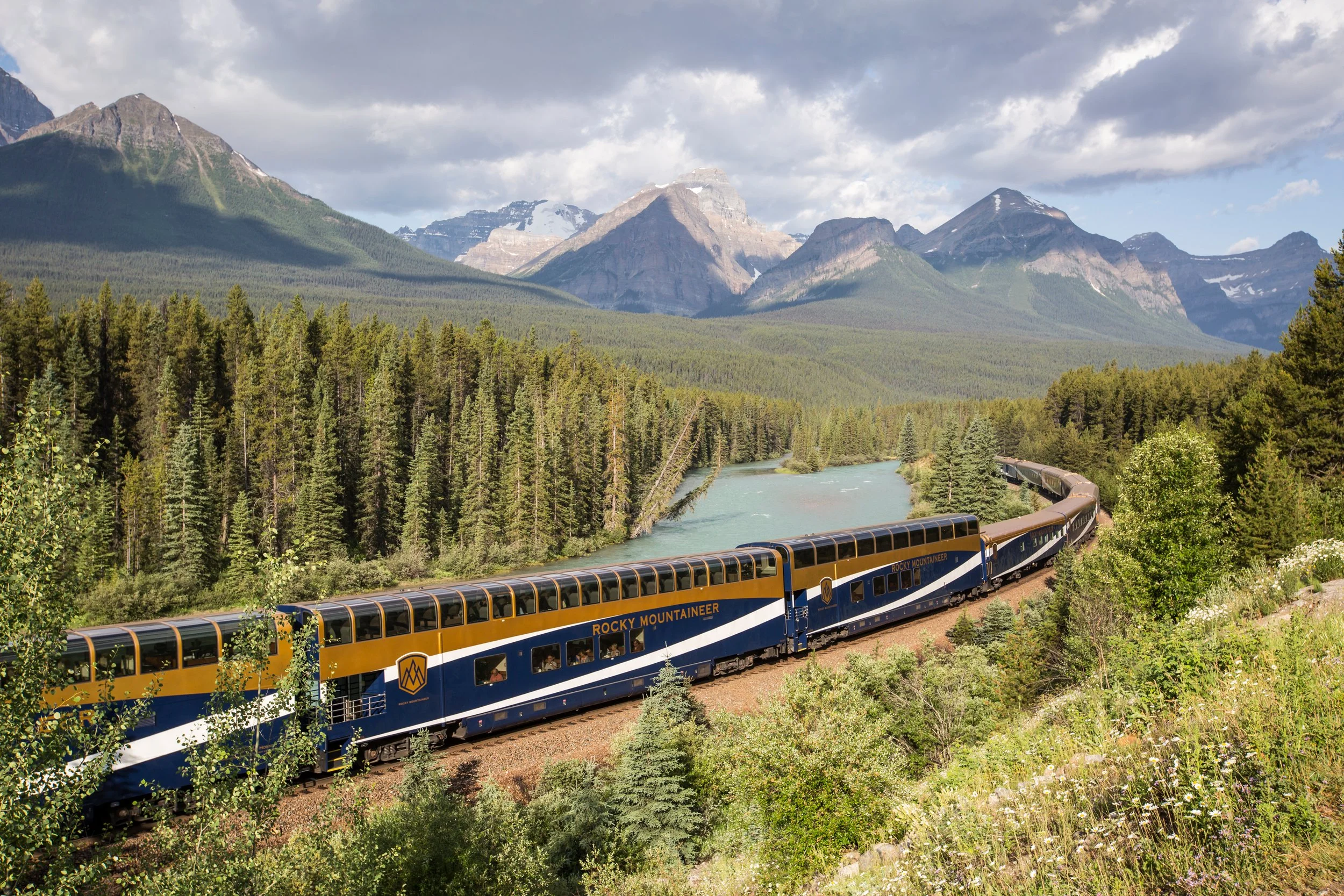 Rocky Mountaineer GoldLeaf train traveling along a river with towering pine forests and the Canadian Rockies in the background.
