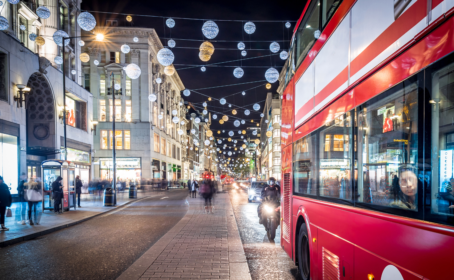 A festive London street at night with sparkling Christmas lights above and a red double-decker bus in the foreground.