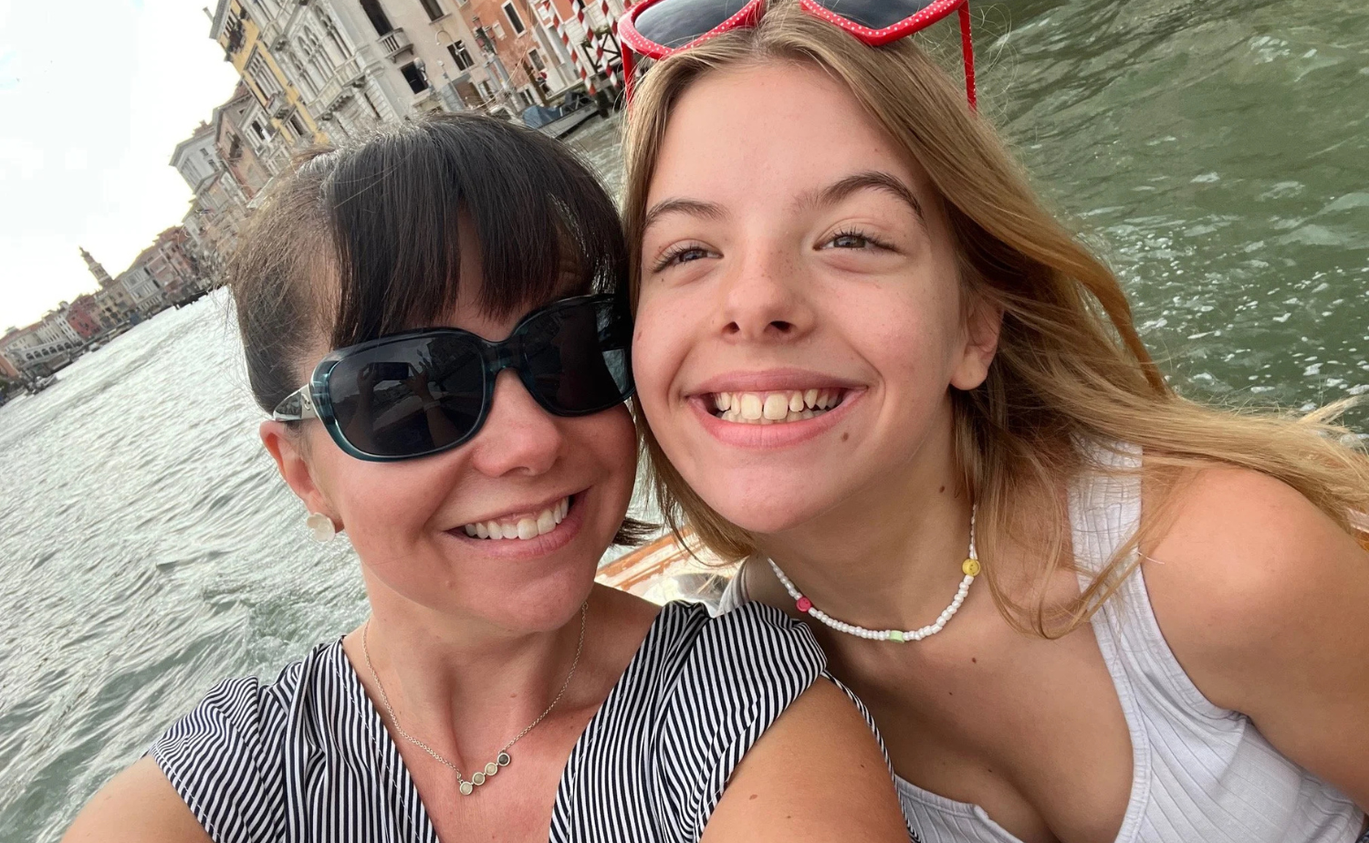 A mother and daughter smiling together on a boat in Venice with canals and historic buildings in the background.