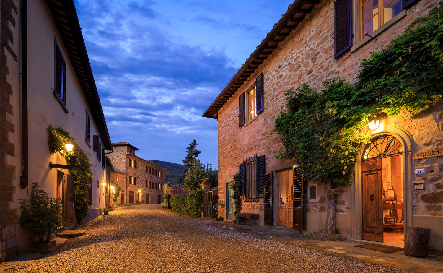 A charming stone village in Tuscany illuminated by warm lights at dusk along a quiet cobblestone street.