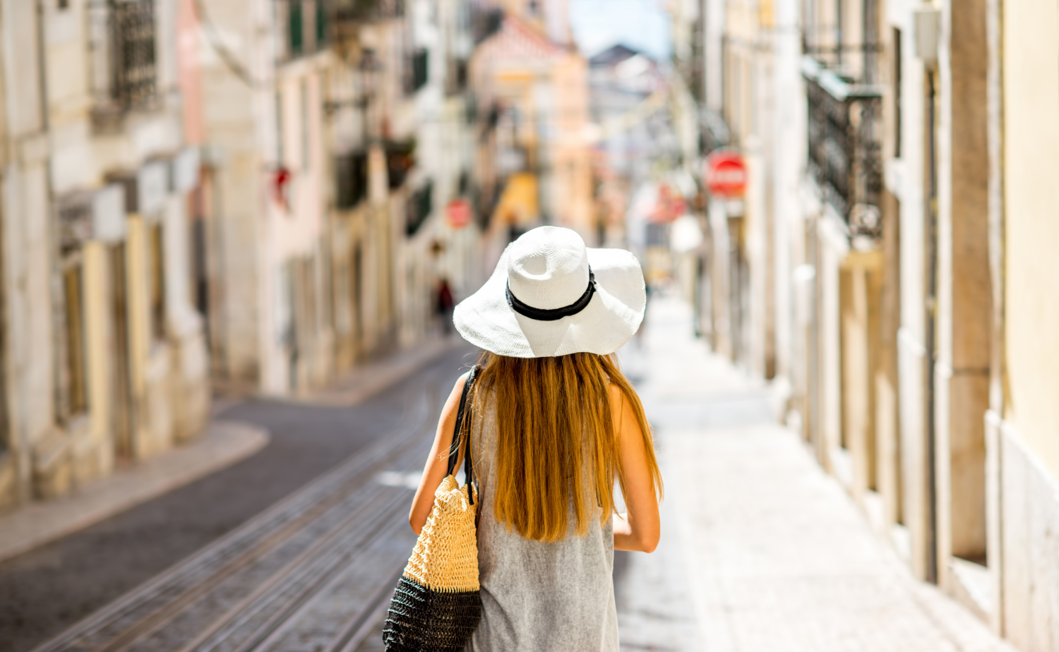 Traveler walking down a European city street with a hat and tote bag, enjoying a relaxed pace.