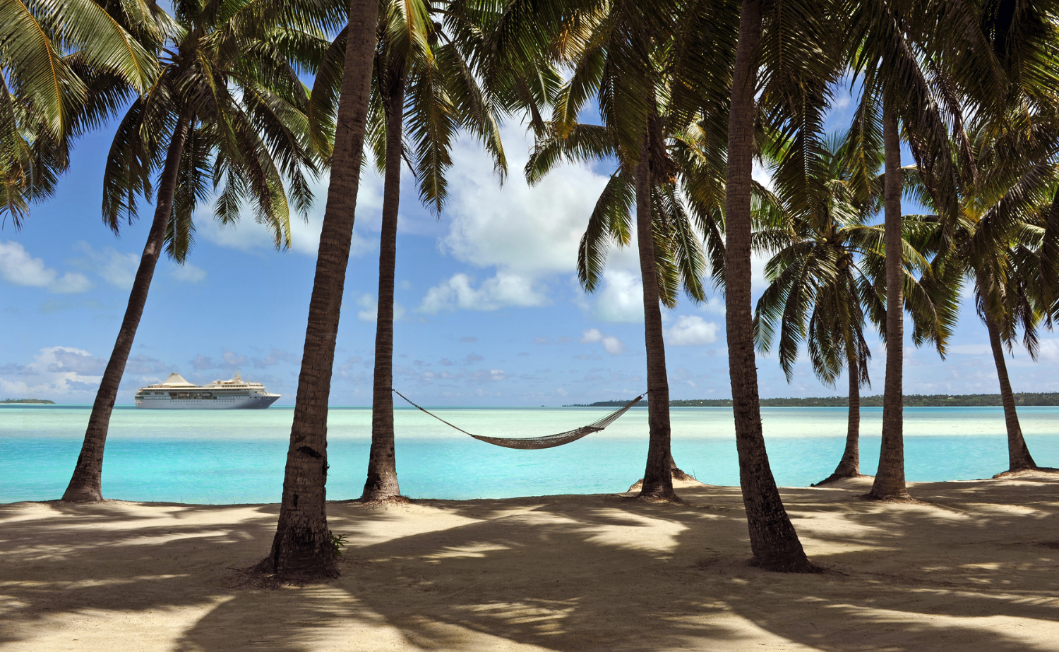 Small cruise ship offshore from a palm-lined beach and turquoise lagoon in French Polynesia.