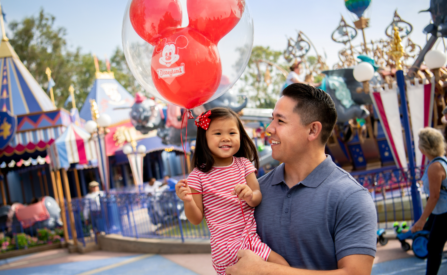 Father holding his young daughter beneath a red Mickey Mouse balloon near Fantasyland attractions at Disneyland.