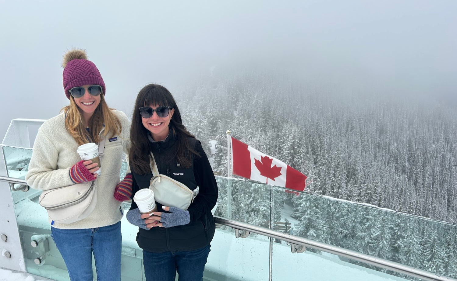 Laura and Molly holding coffee on the snowy Sulphur Mountain viewing platform with the Canadian wilderness behind them.