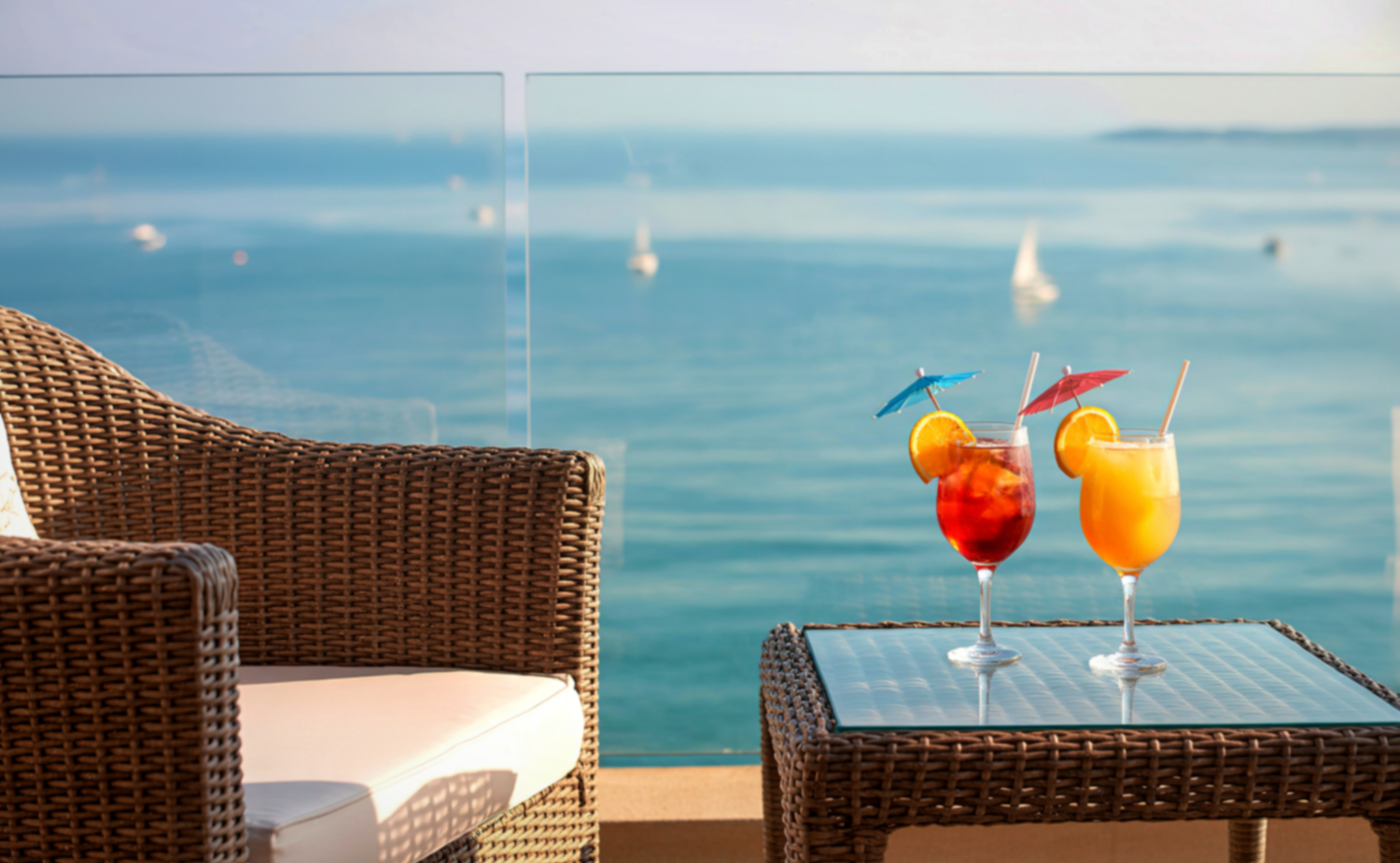 Two colorful tropical drinks on a wicker table overlooking calm blue ocean waters with sailboats in the distance.