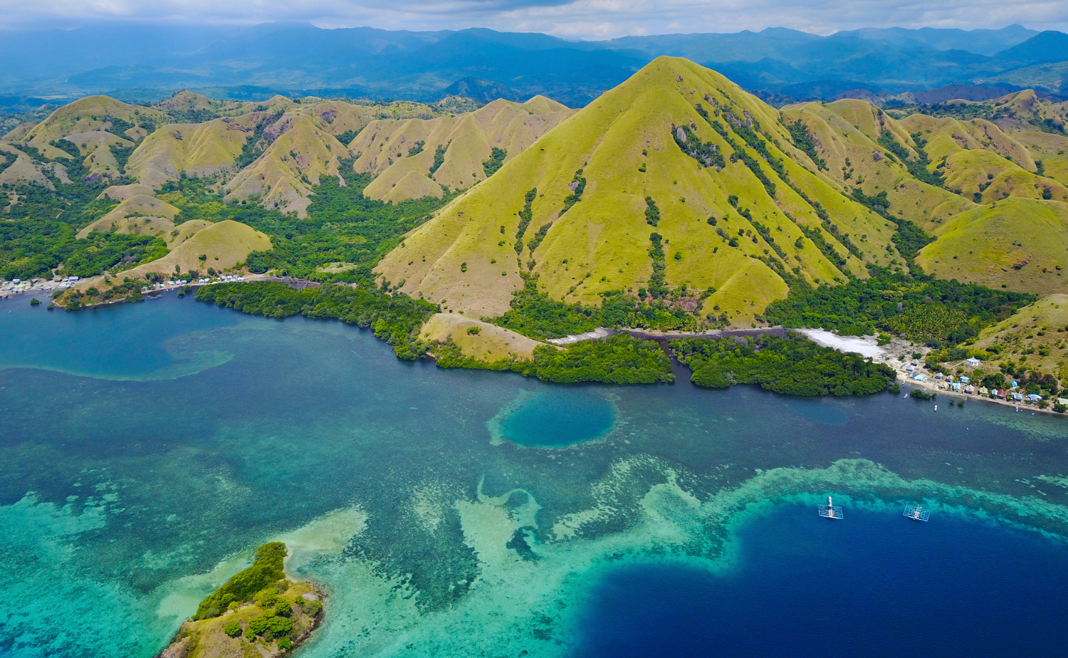 Rolling green island hills meeting a turquoise lagoon with coral reefs visible beneath clear water.