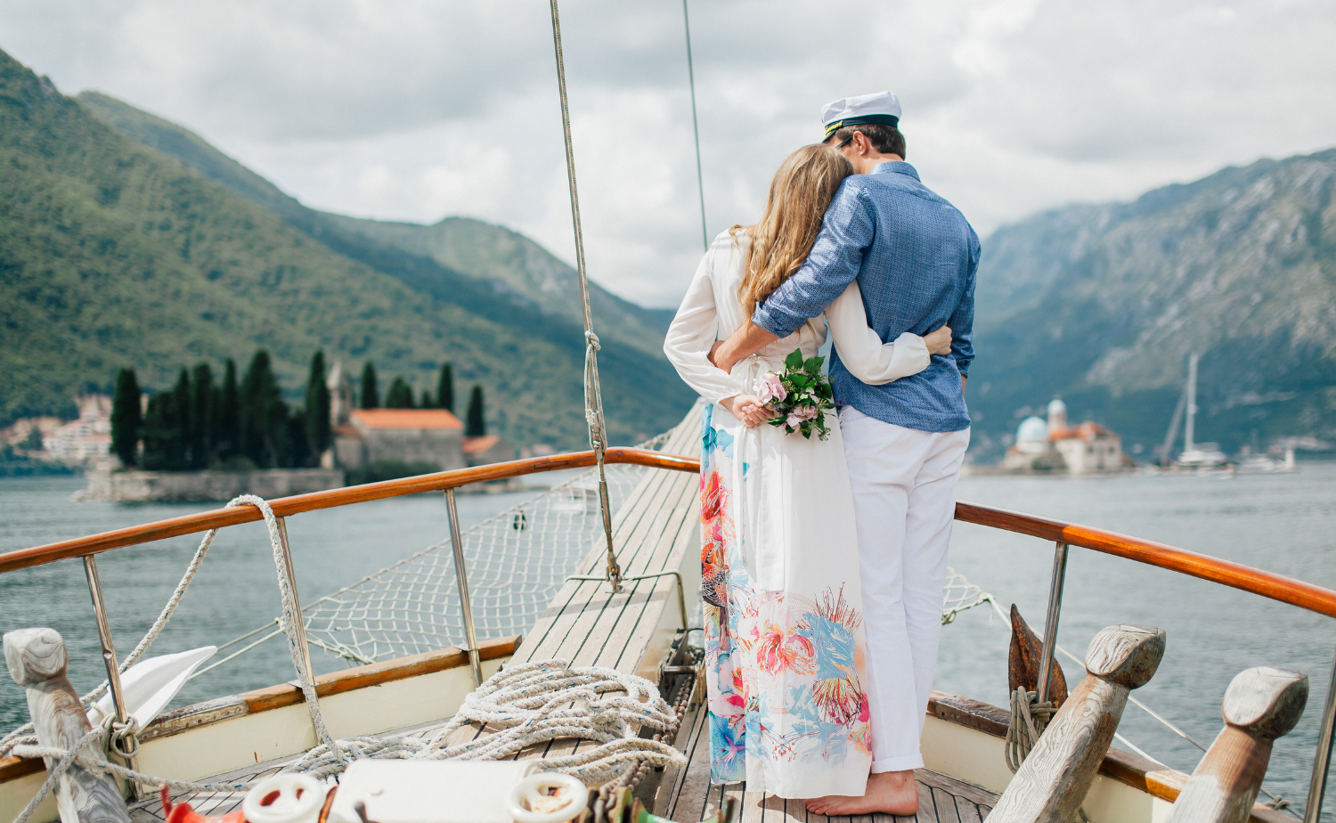 A couple stands together on the deck of a sailboat, surrounded by mountains and calm blue water, capturing a romantic travel moment